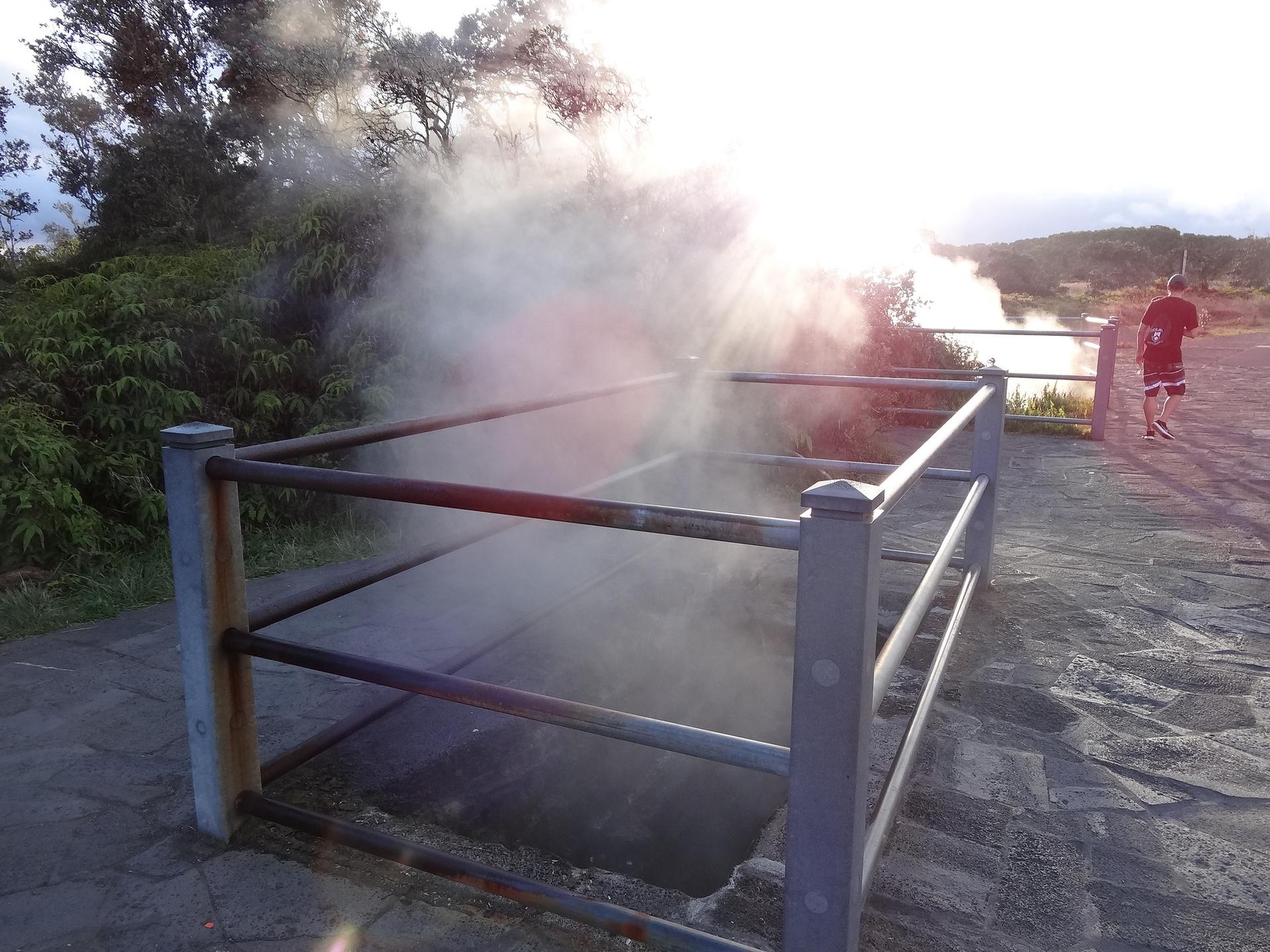 A steaming ground vent with a metal railing, person in red in sunlight.