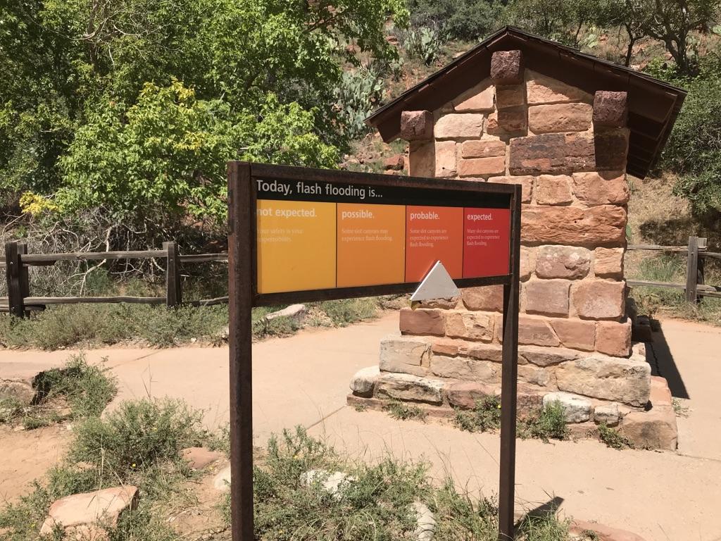 A flash flood warning sign stands next to a stone building and green trees.