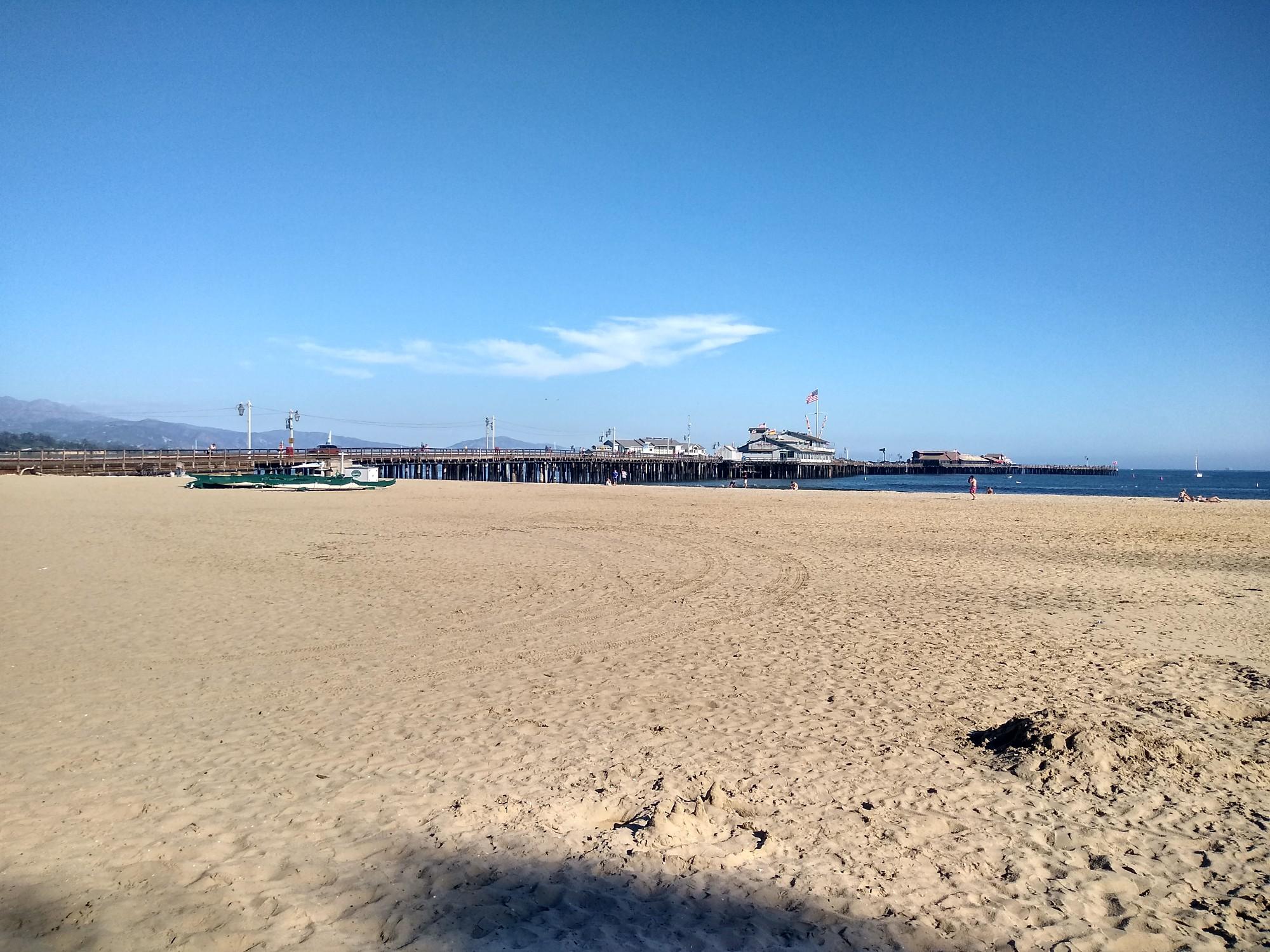 Expansive sandy beach with a long pier and boats in the distance, sunny.