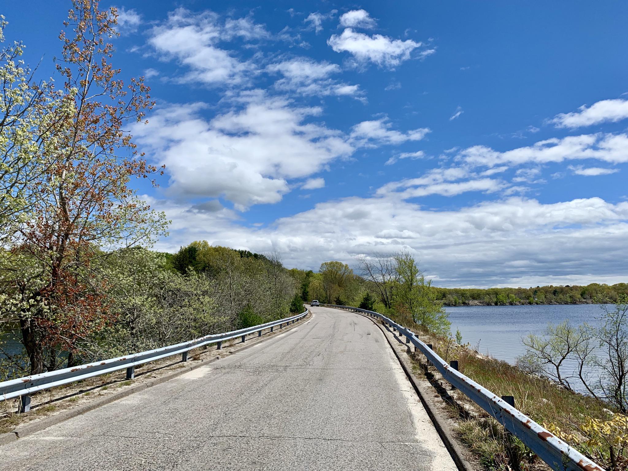 A winding road with guardrails next to a lake under a bright sky.