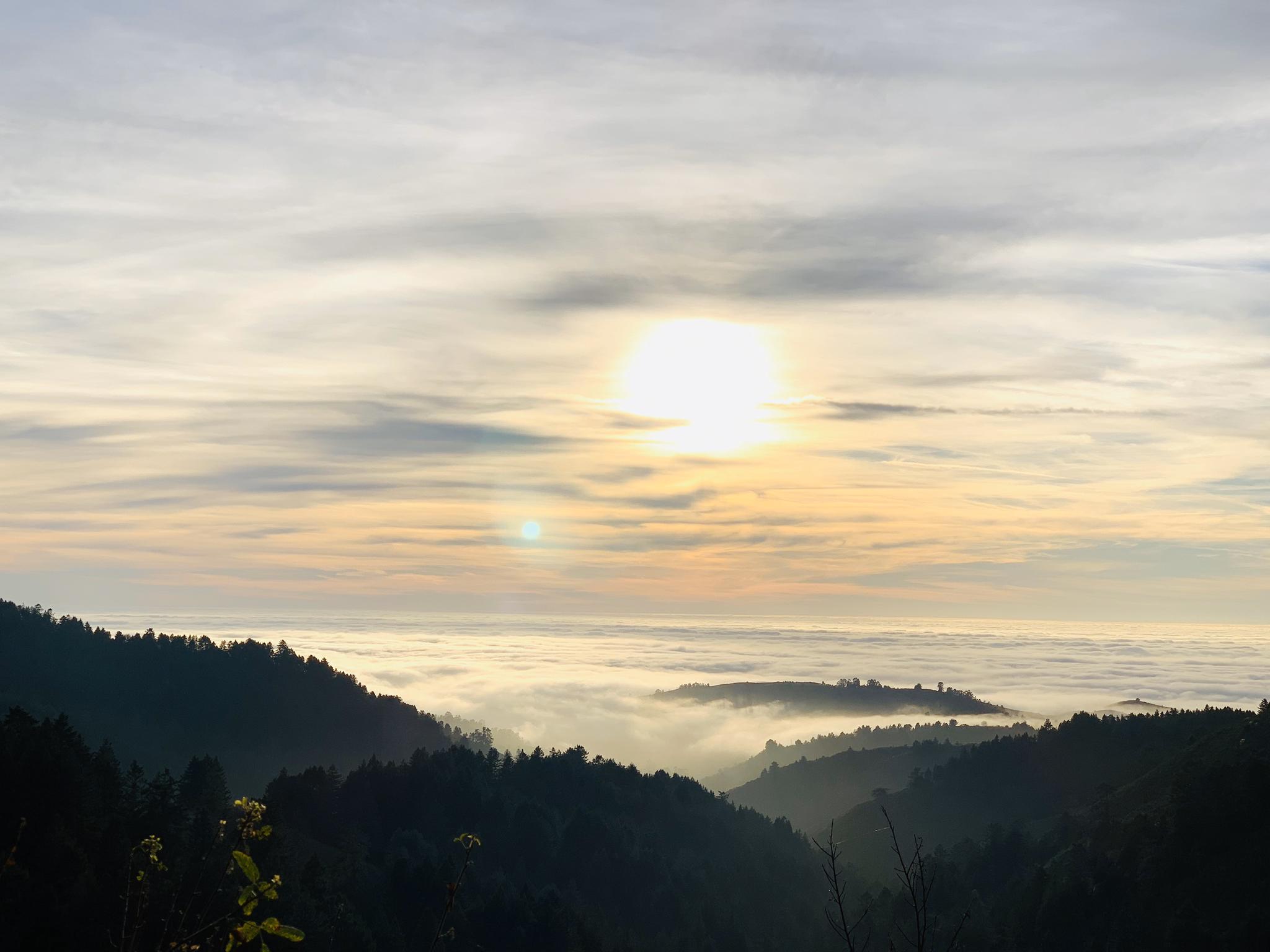 Bright sun setting over a dramatic cloud inversion above forested hills.