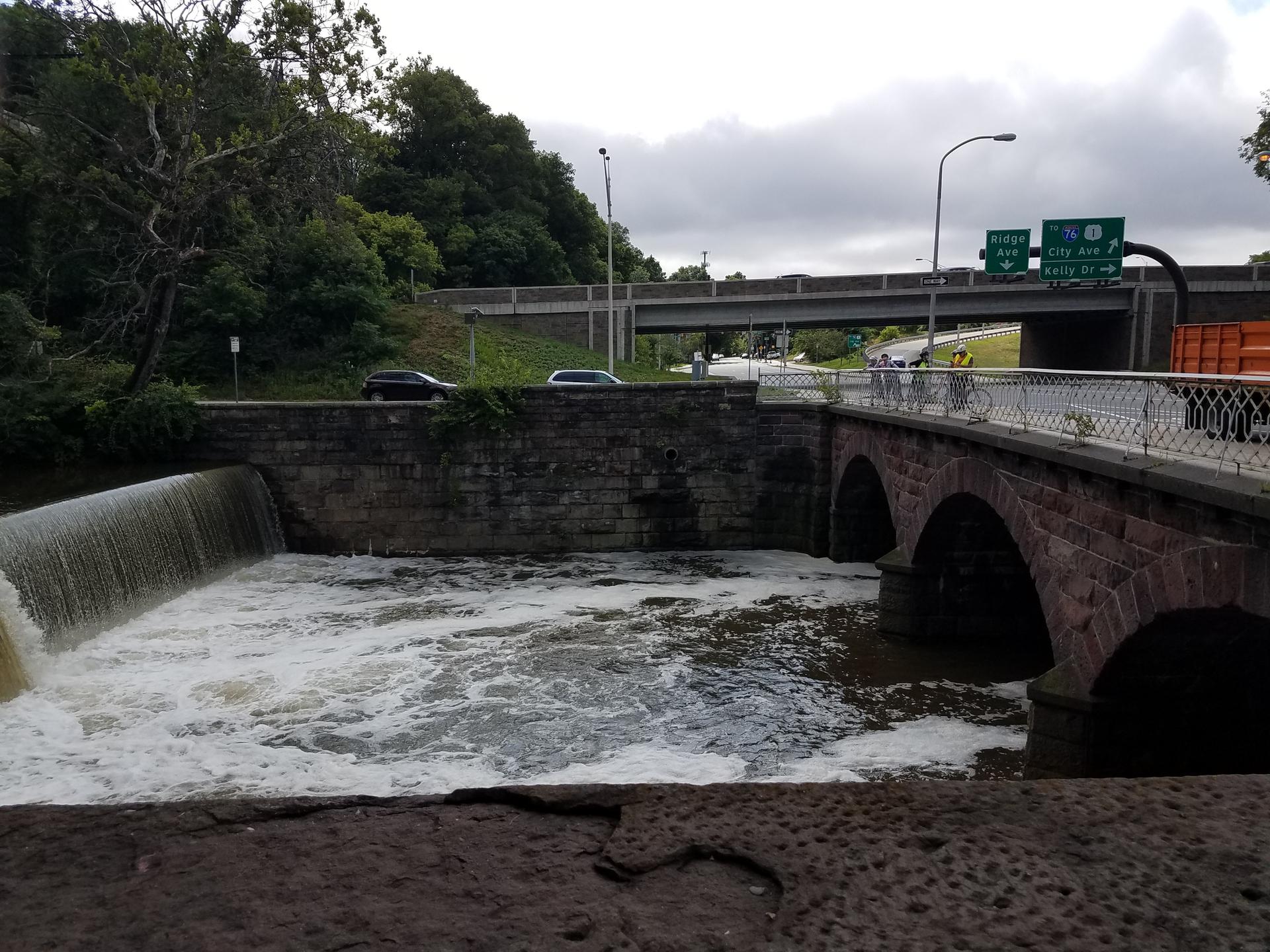 Stone bridge over dam with rushing water, highway overpass, overcast day.