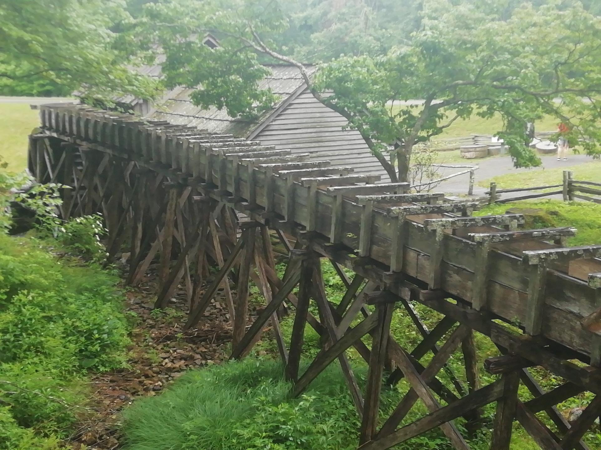 Long wooden aqueduct on stilts, winding through green foliage with a building behind.