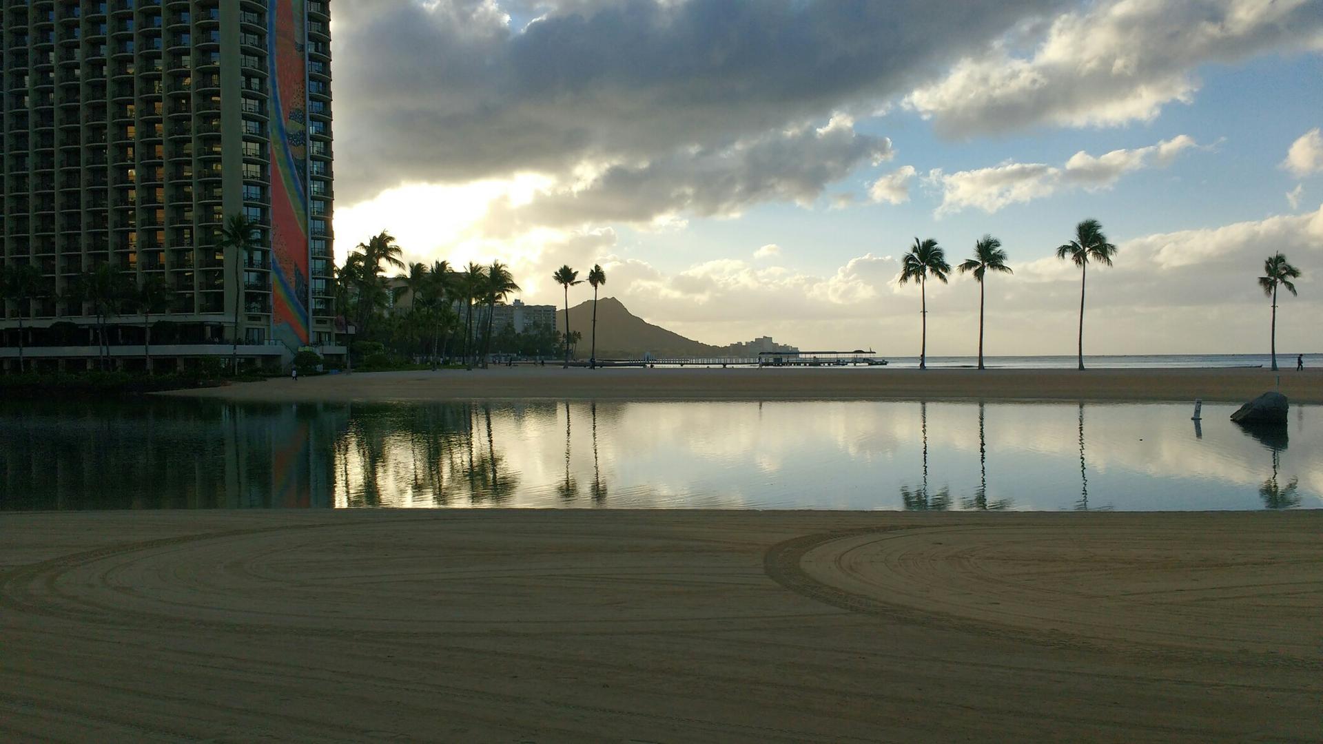 Calm water reflects palm trees and a distant mountain at sunset, next to a large building.