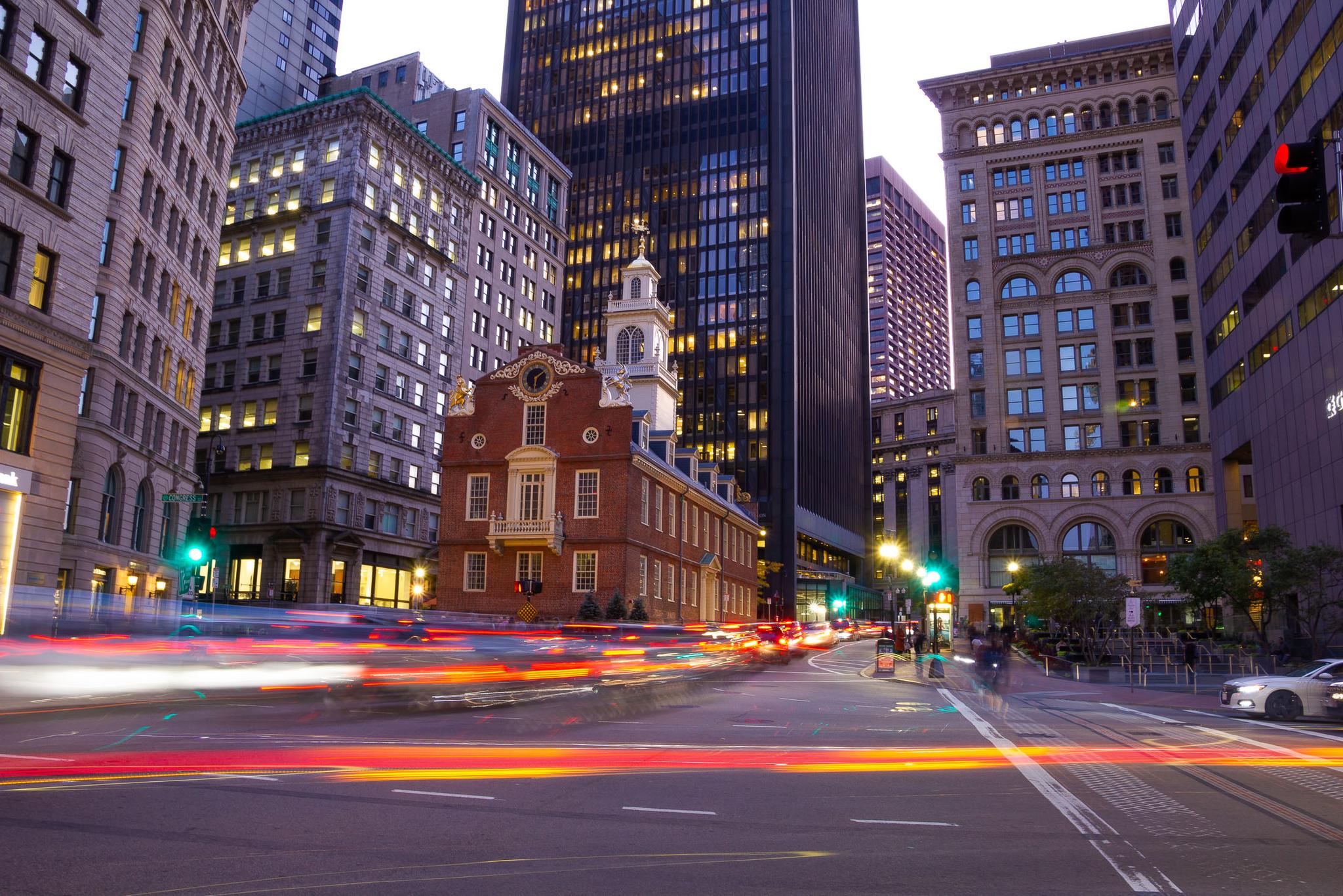 Historic brick building amidst modern skyscrapers with vibrant car light trails at dusk.