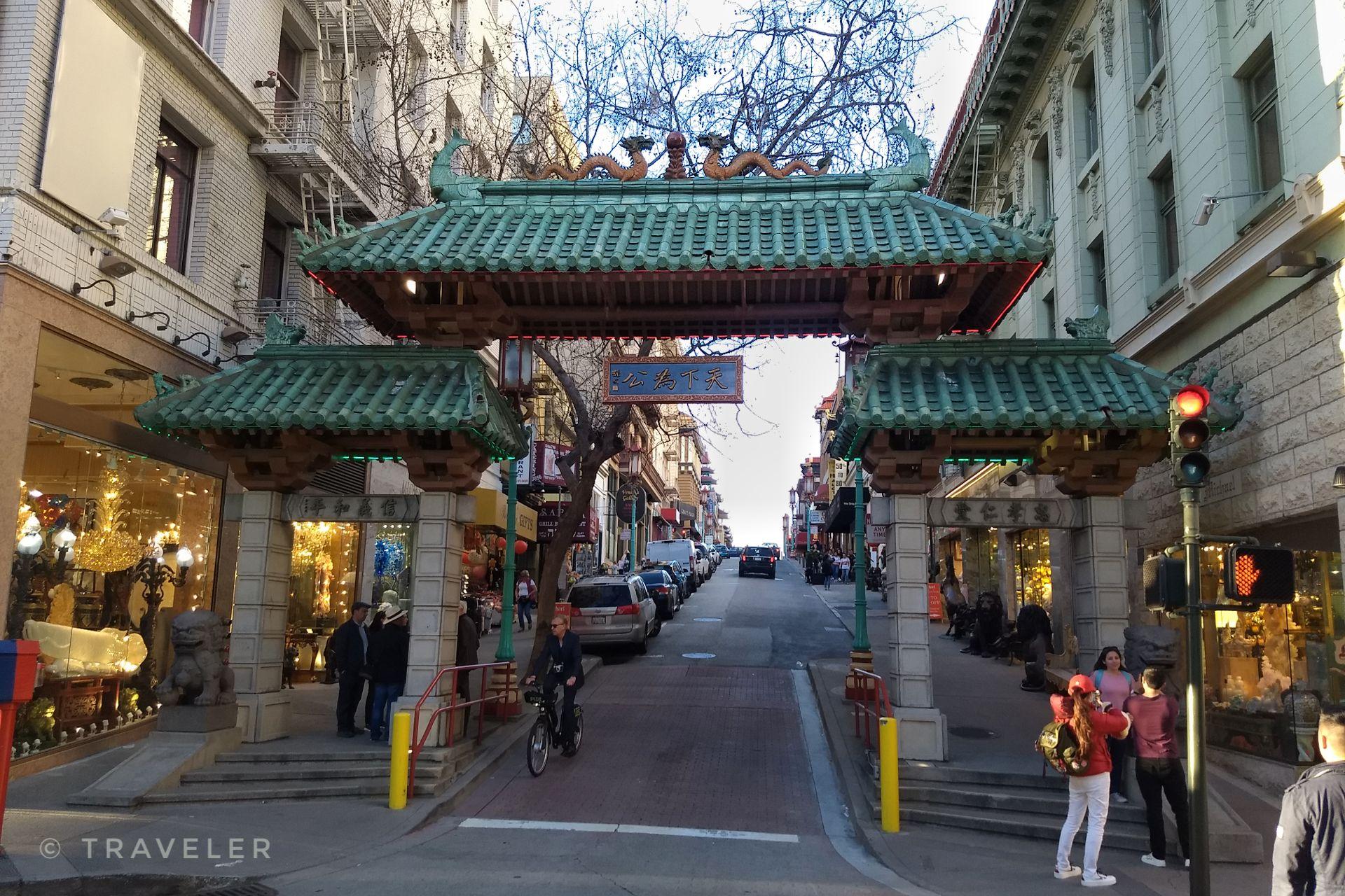 Ornate archway over a city street with buildings, cars, and pedestrians.