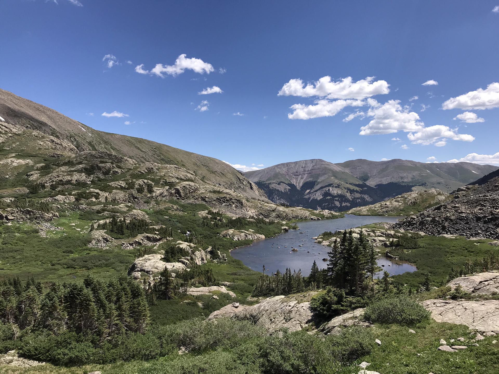 A scenic mountain lake nestled in a green valley with distant peaks under a blue sky.