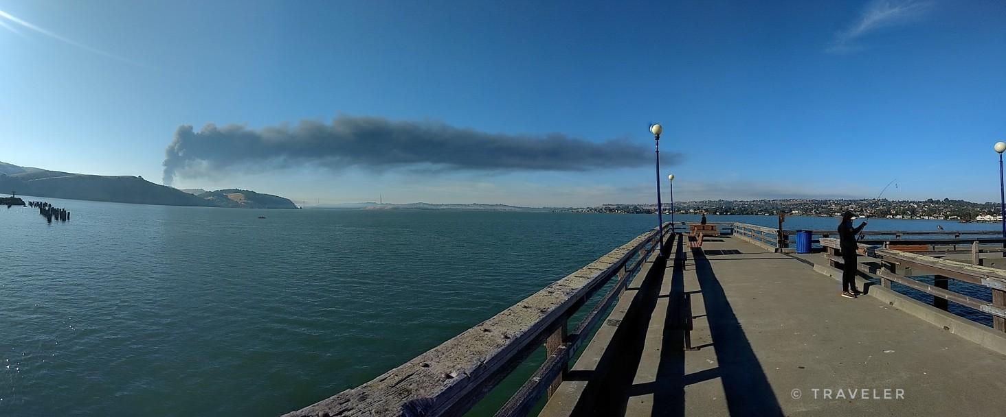 A person stands on a long pier over water with a distant smoke plume.