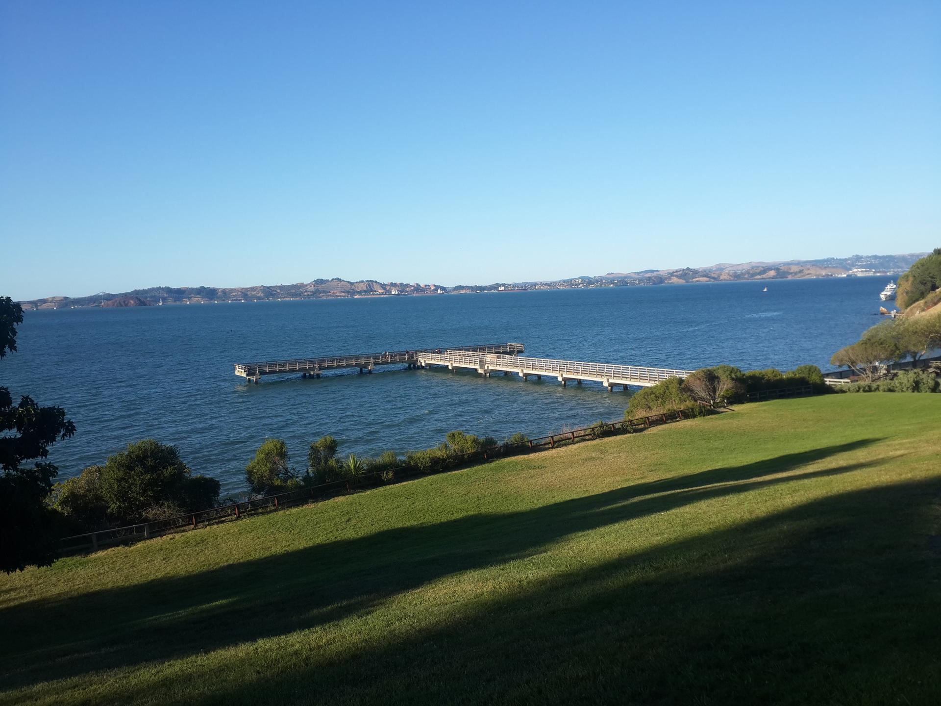 Long pier extending into blue water, green grass, and distant hills on a sunny day.