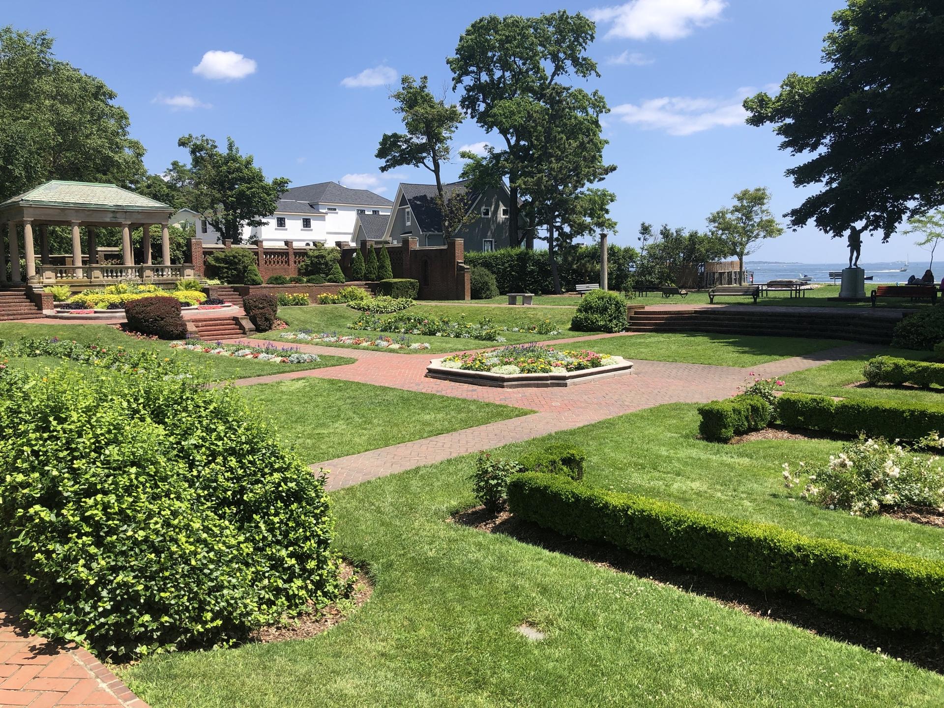 Sunny formal garden with brick paths, green lawns, hedges, and a gazebo.