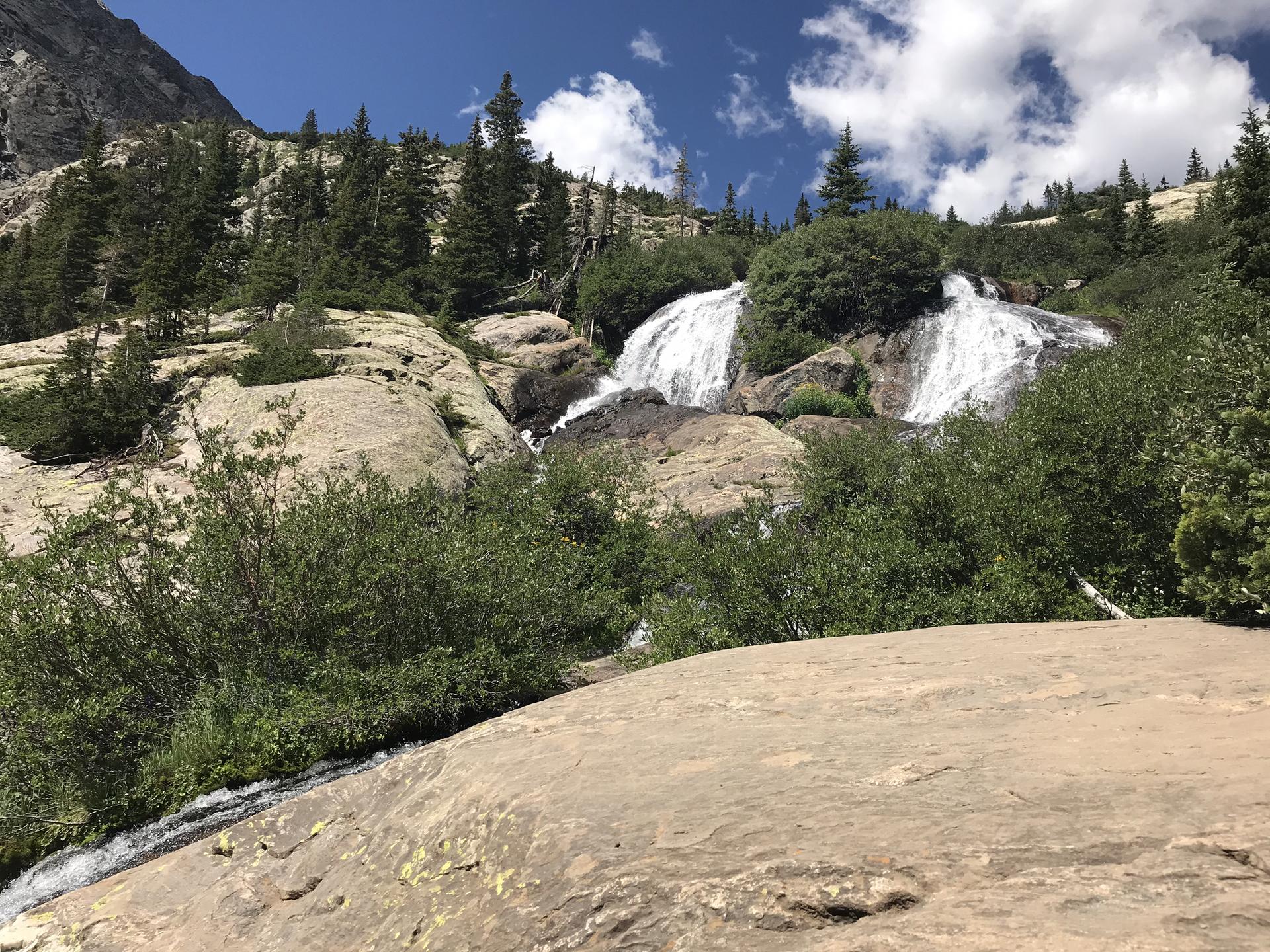 A multi-tiered waterfall cascades over large rocks, surrounded by green trees.