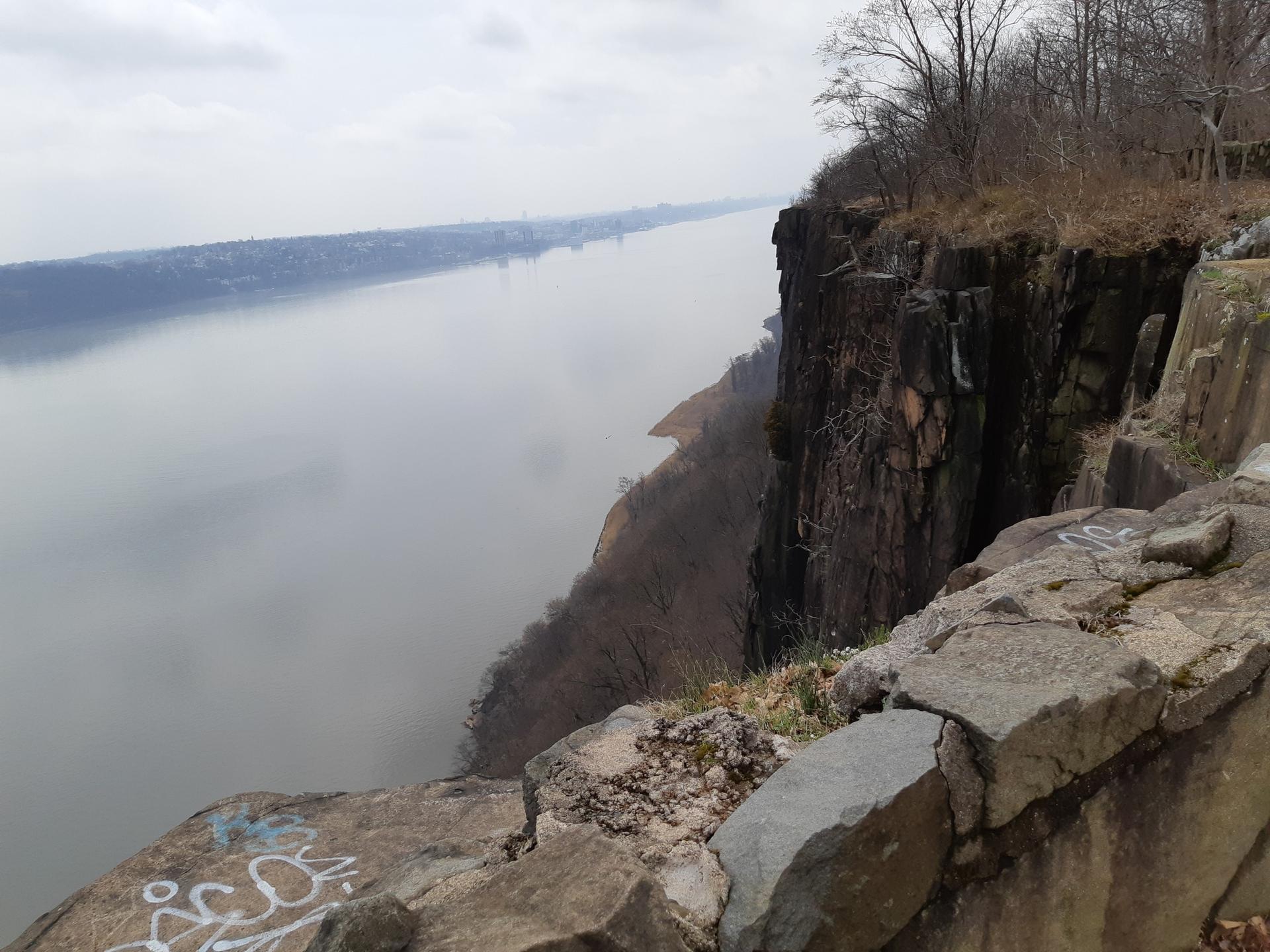 Overcast view of a wide river next to a tall, rocky cliff.