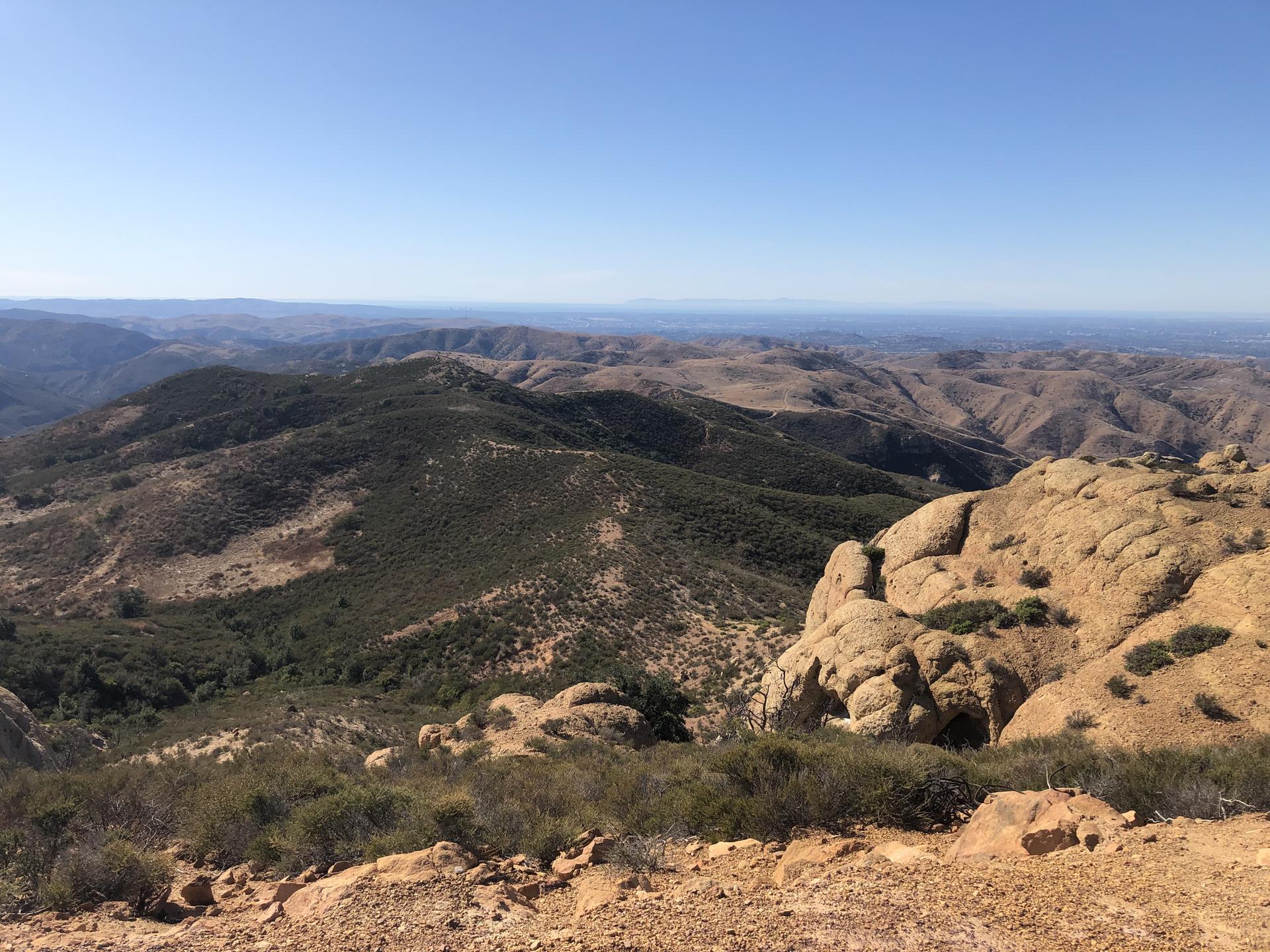 Vast mountain landscape with rocky outcrops and green hills under a clear blue sky.