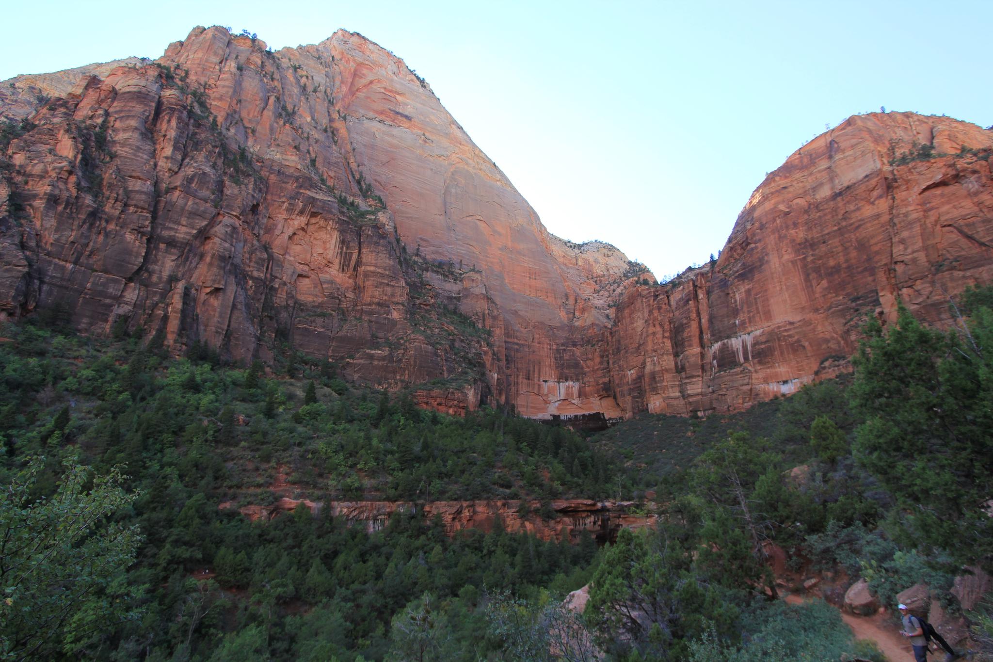 Towering red rock mountain face with green trees at its base under soft light.