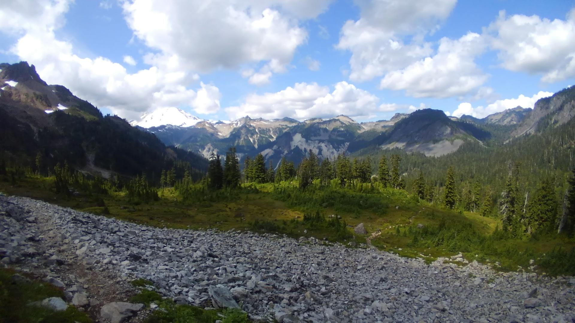 Rocky foreground, green valley, and distant snow-capped mountains under a cloudy sky.