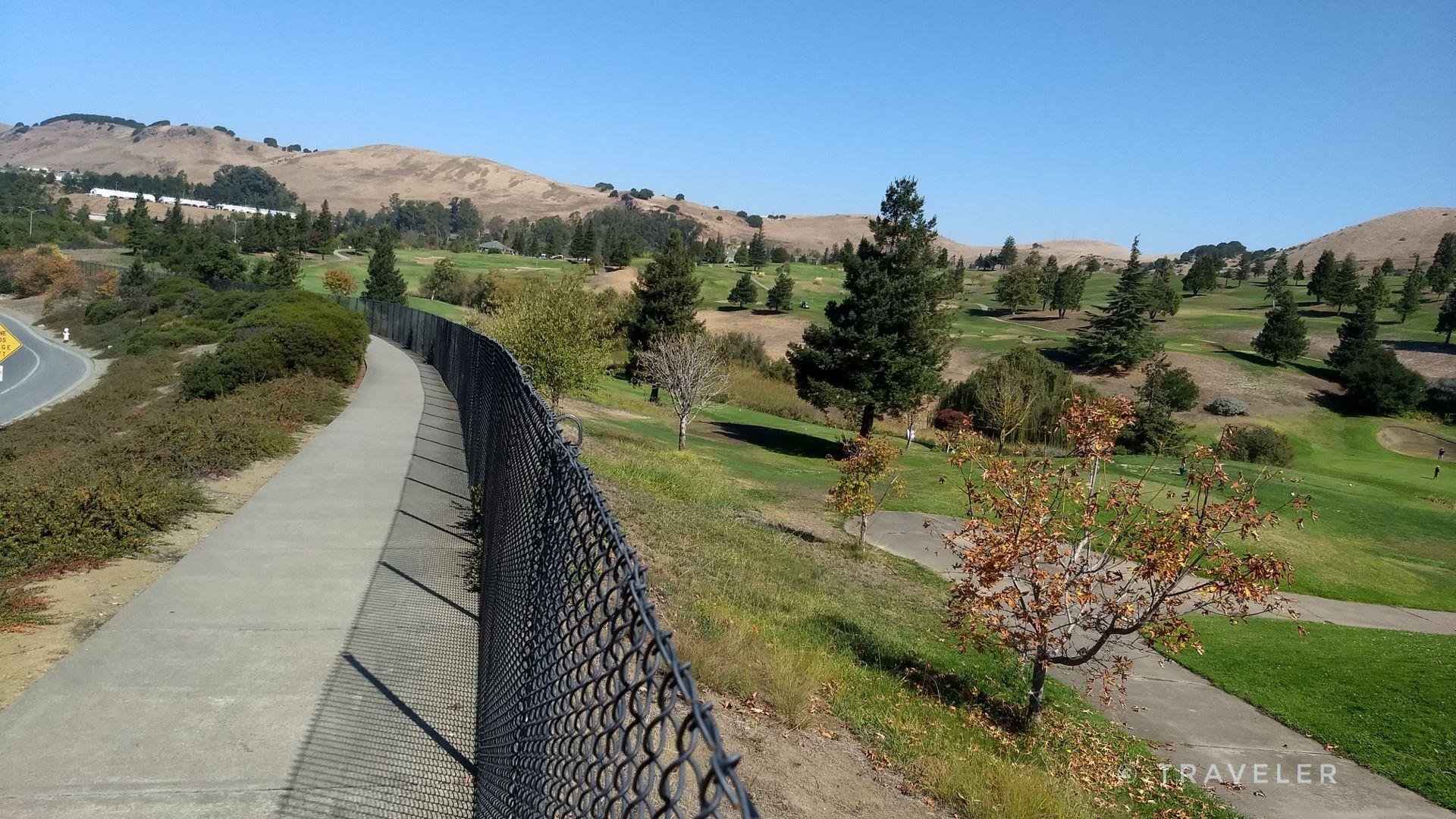 Paved path alongside a chain-link fence, overlooking a green golf course.