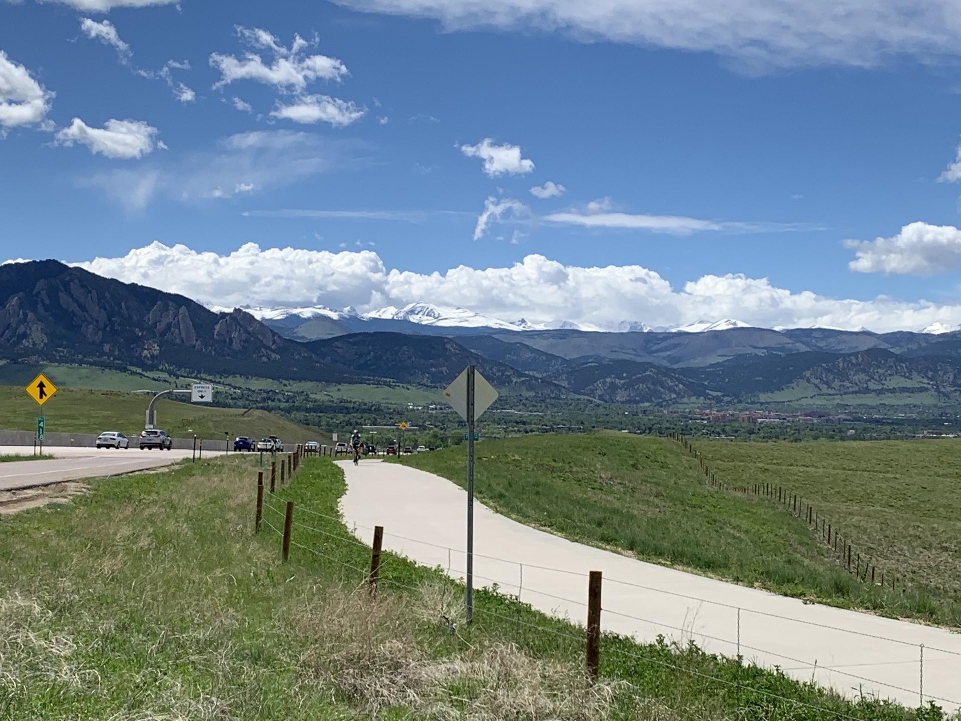 Paved path winding through green hills with distant mountains under a cloudy sky.