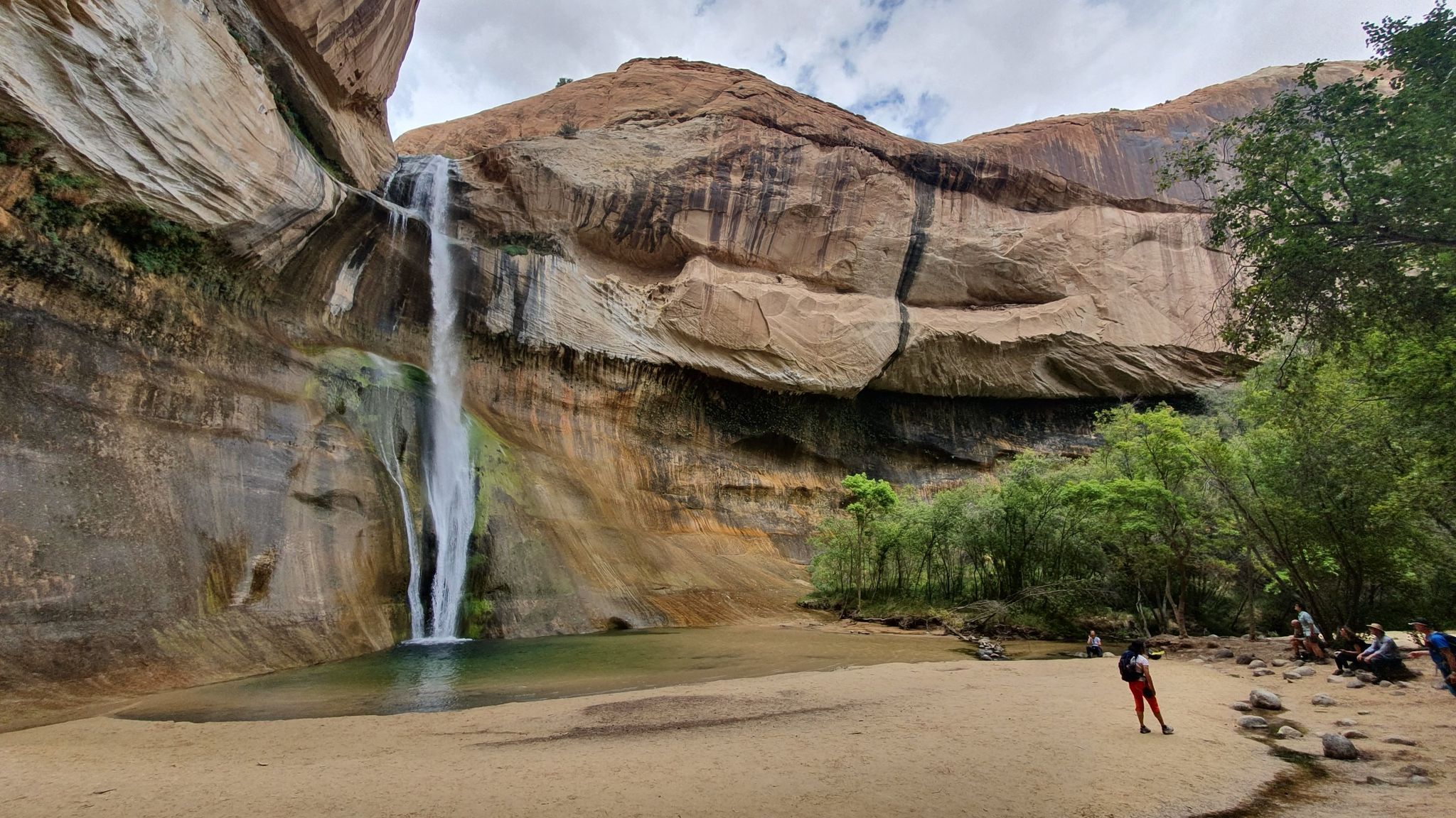 A tall waterfall plunges into a pool, with people on a sandy bank. - Lower Calf Creek Falls Trail