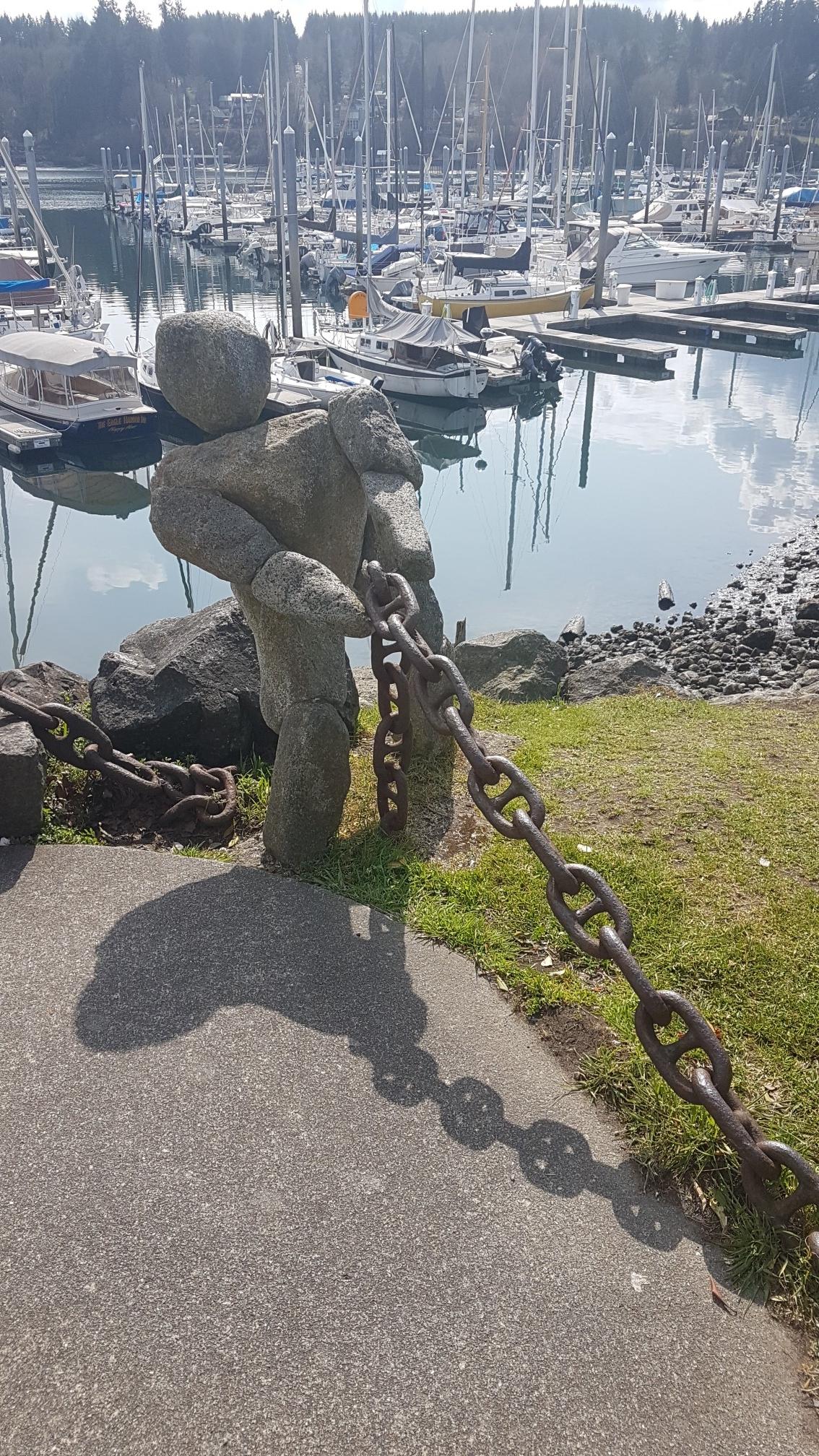 Stone statue pulling a large chain beside a sunny marina with boats.
