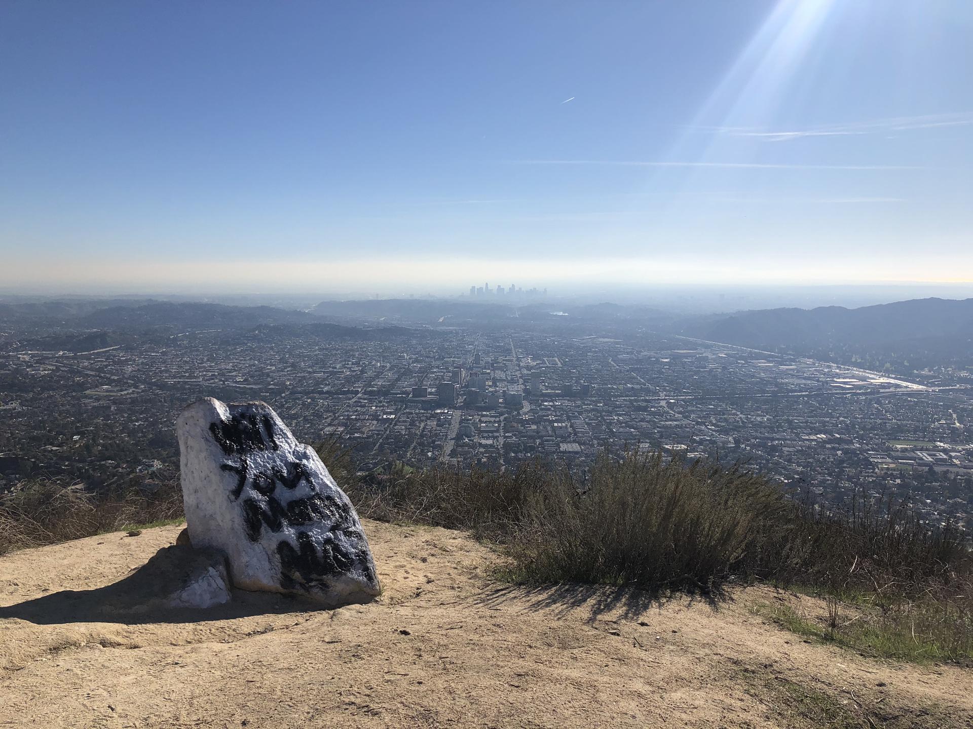 Large white rock on a hilltop overlooking a sprawling city under bright sun.