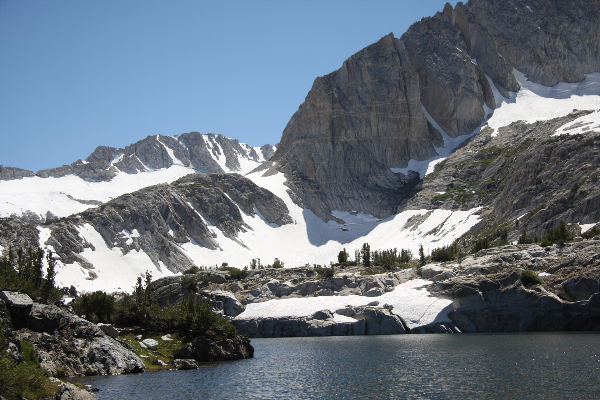 Dark lake at the base of snow-covered, rugged mountains on a sunny day.