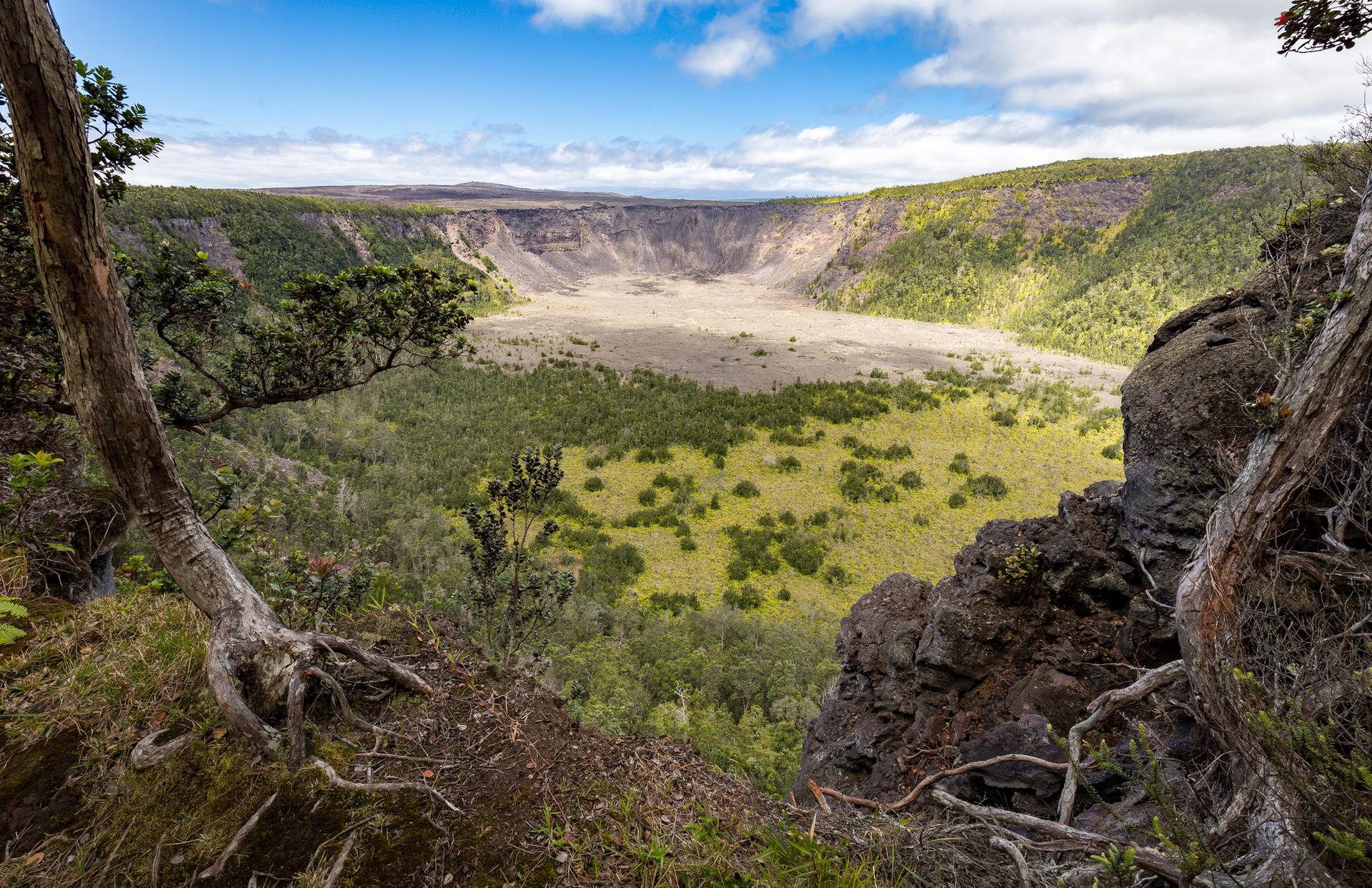 Wide view of a large volcanic crater with green vegetation under a bright sky.