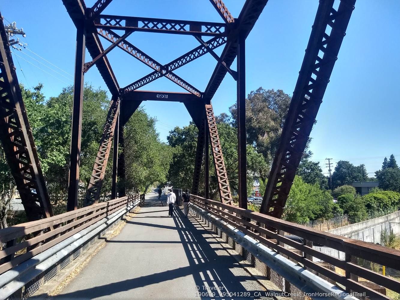 People walking on a metal truss bridge over a paved path on a sunny day. - Sunol Depot loop
