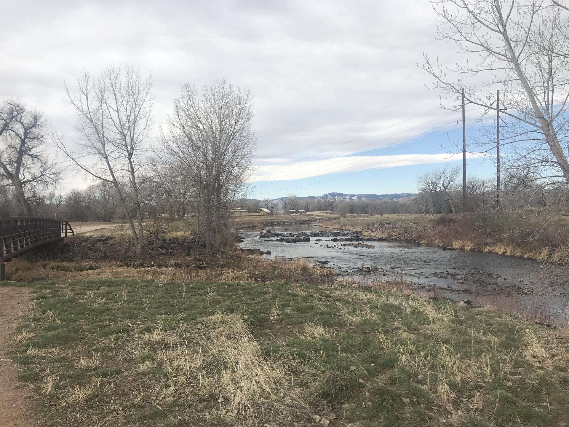 River with rocks, bare trees, and a bridge under a cloudy sky.