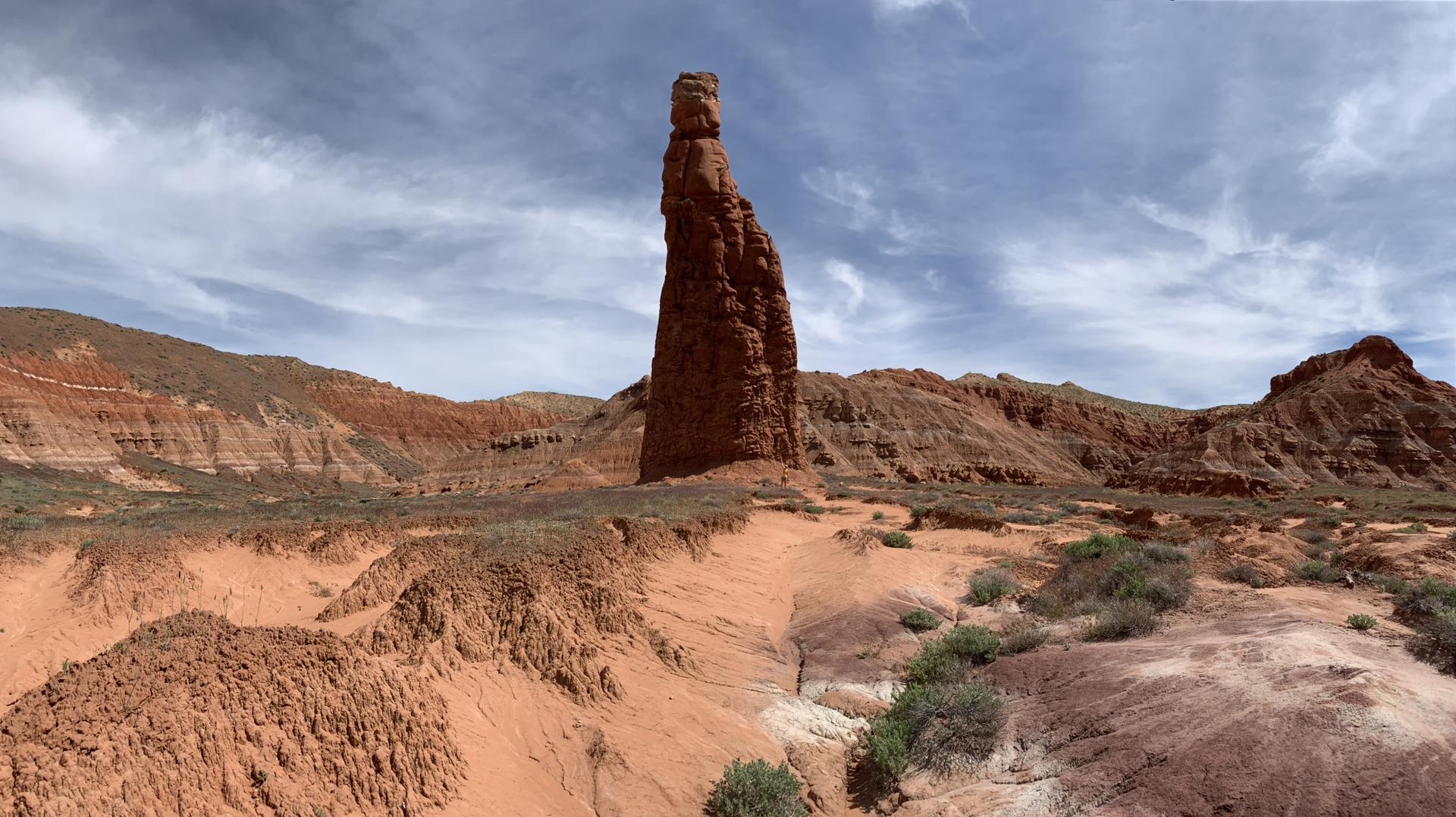 A tall, slender red rock formation stands prominently in an arid landscape.