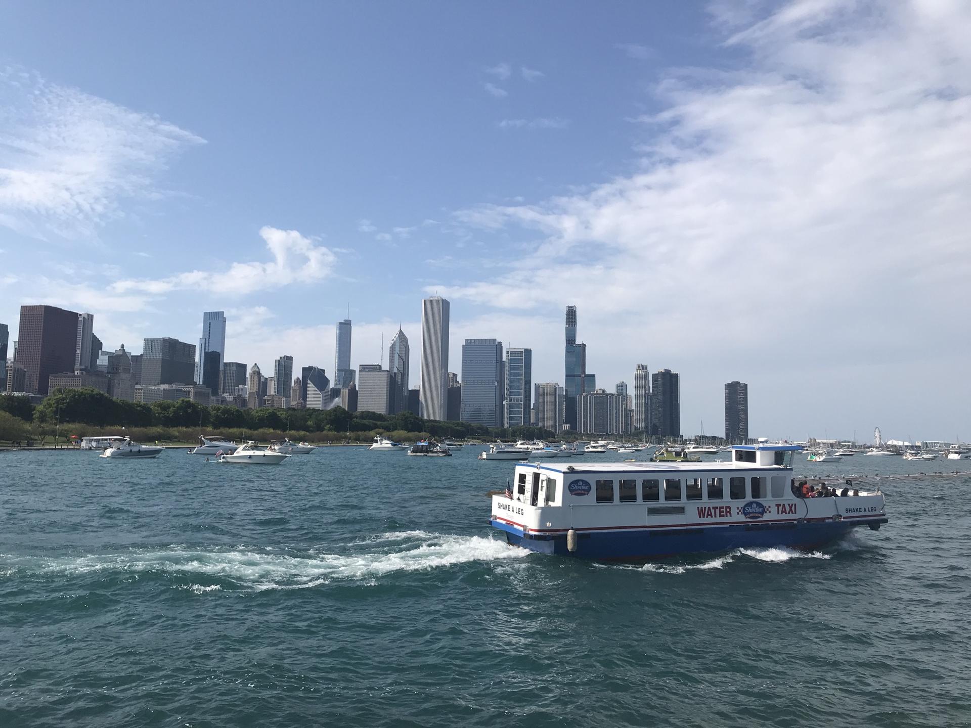 A water taxi cruises past a distant city skyline on a sunny day.