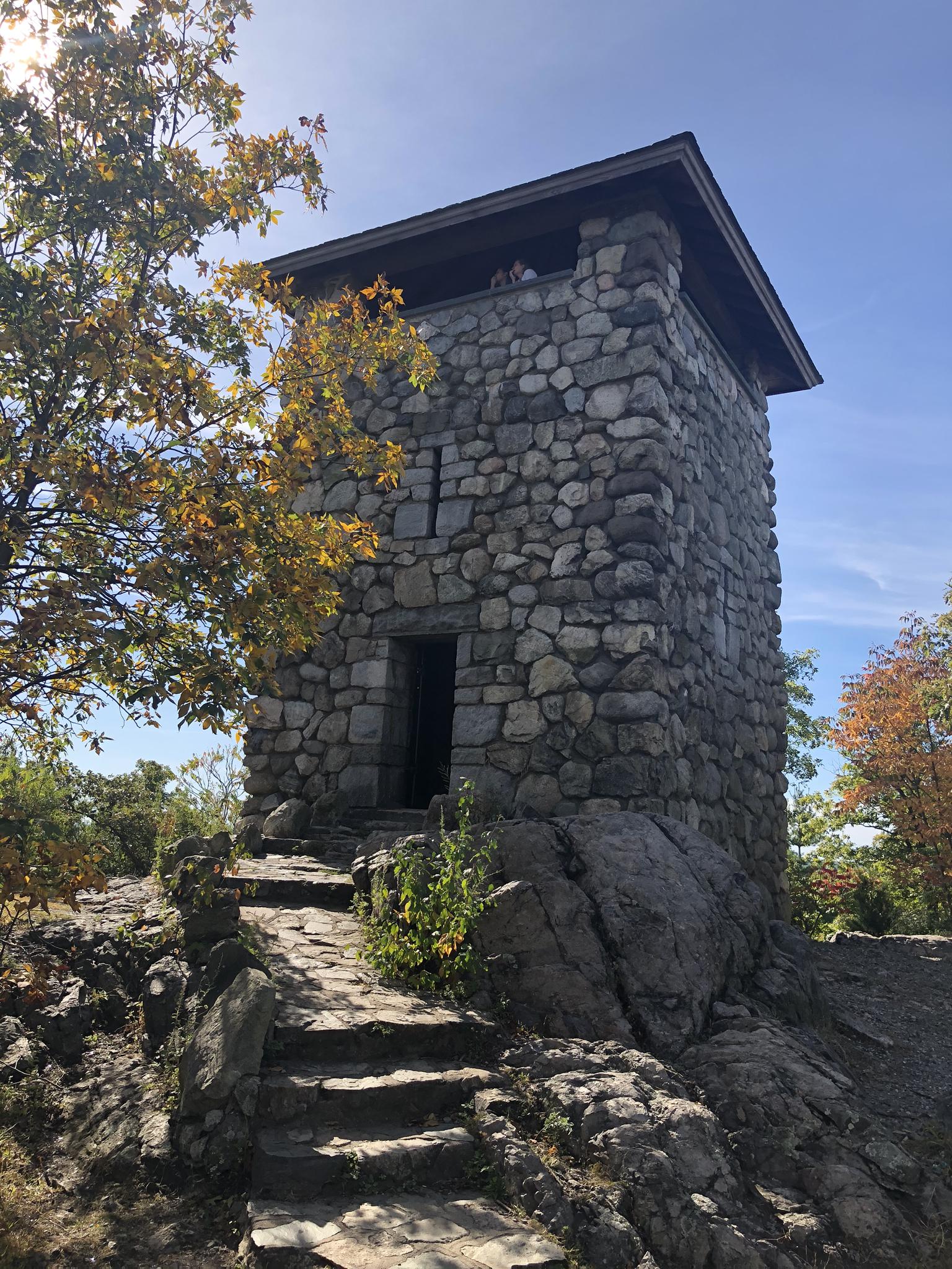 Historic stone tower on a rocky hill, surrounded by autumn trees, bright sun.