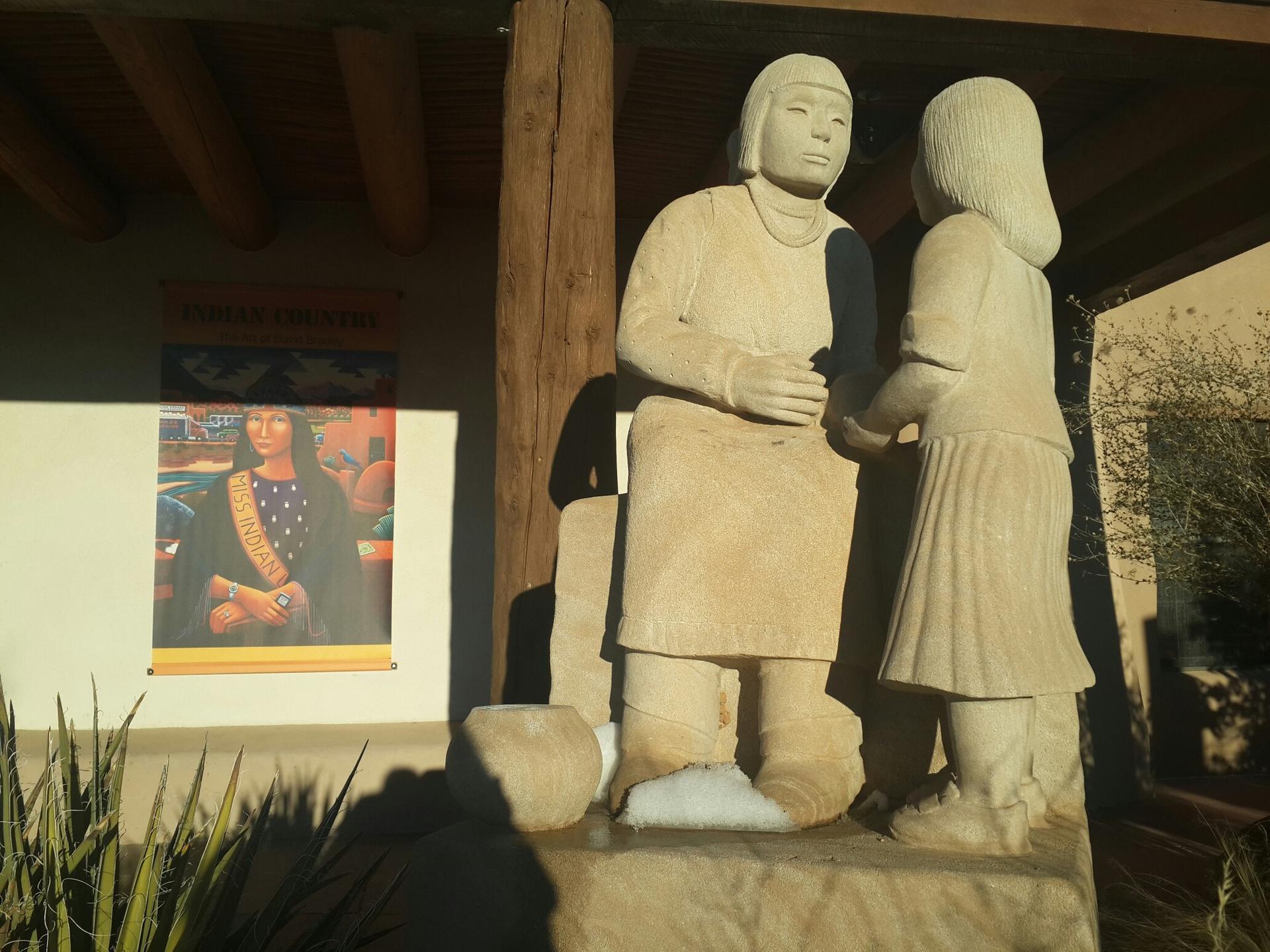 Stone statues of an adult and child under a wooden beam, bright sunlight.