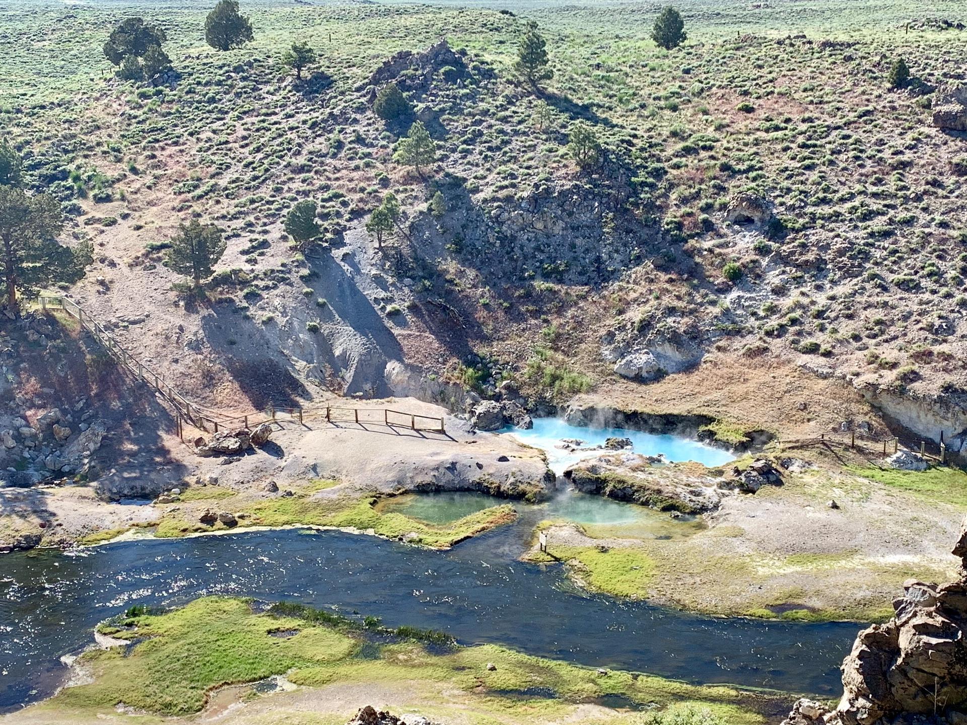 Aerial view of a river, green banks, and bright blue hot springs.