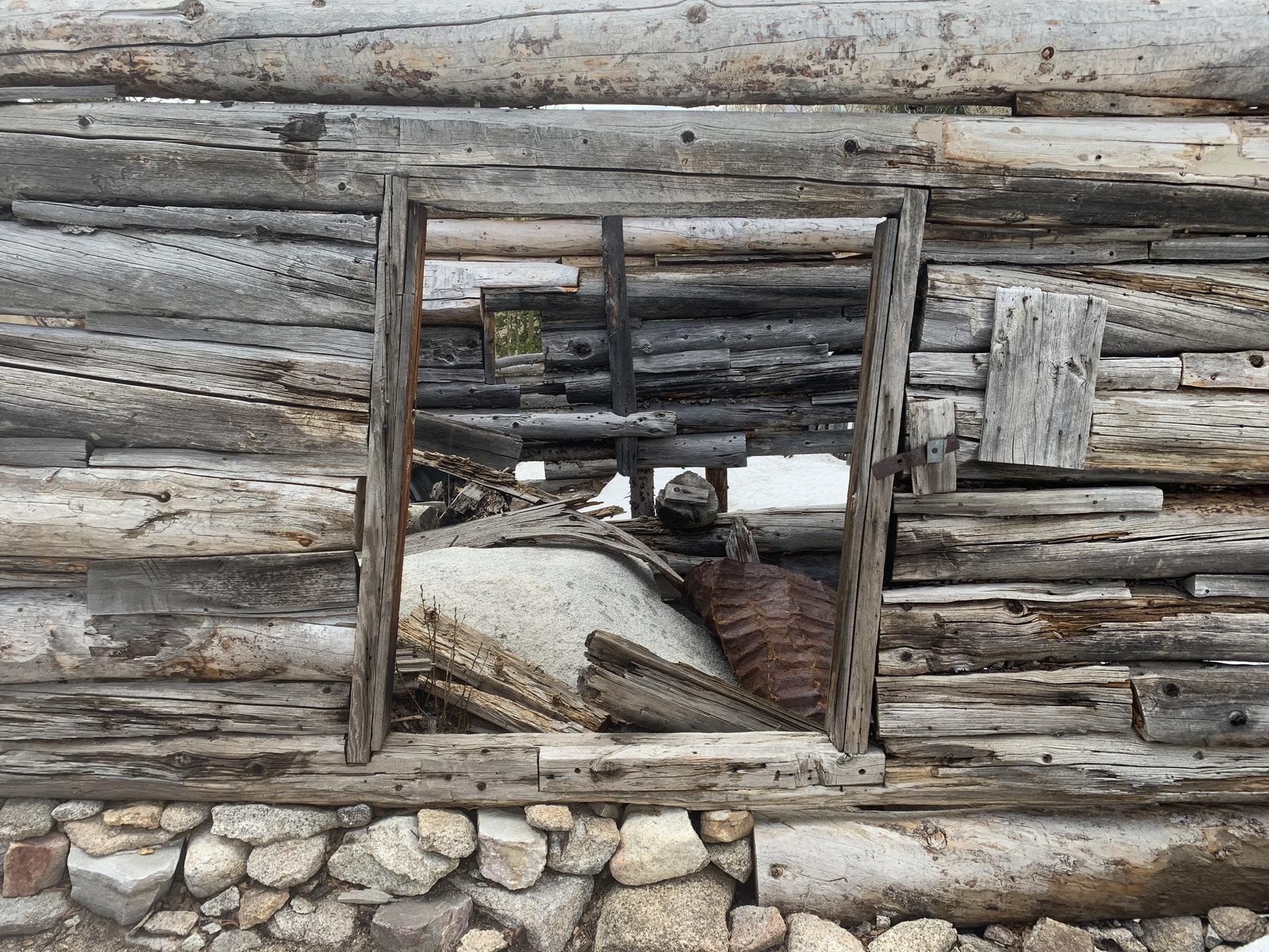 Weathered log cabin wall with a window opening and stone foundation.