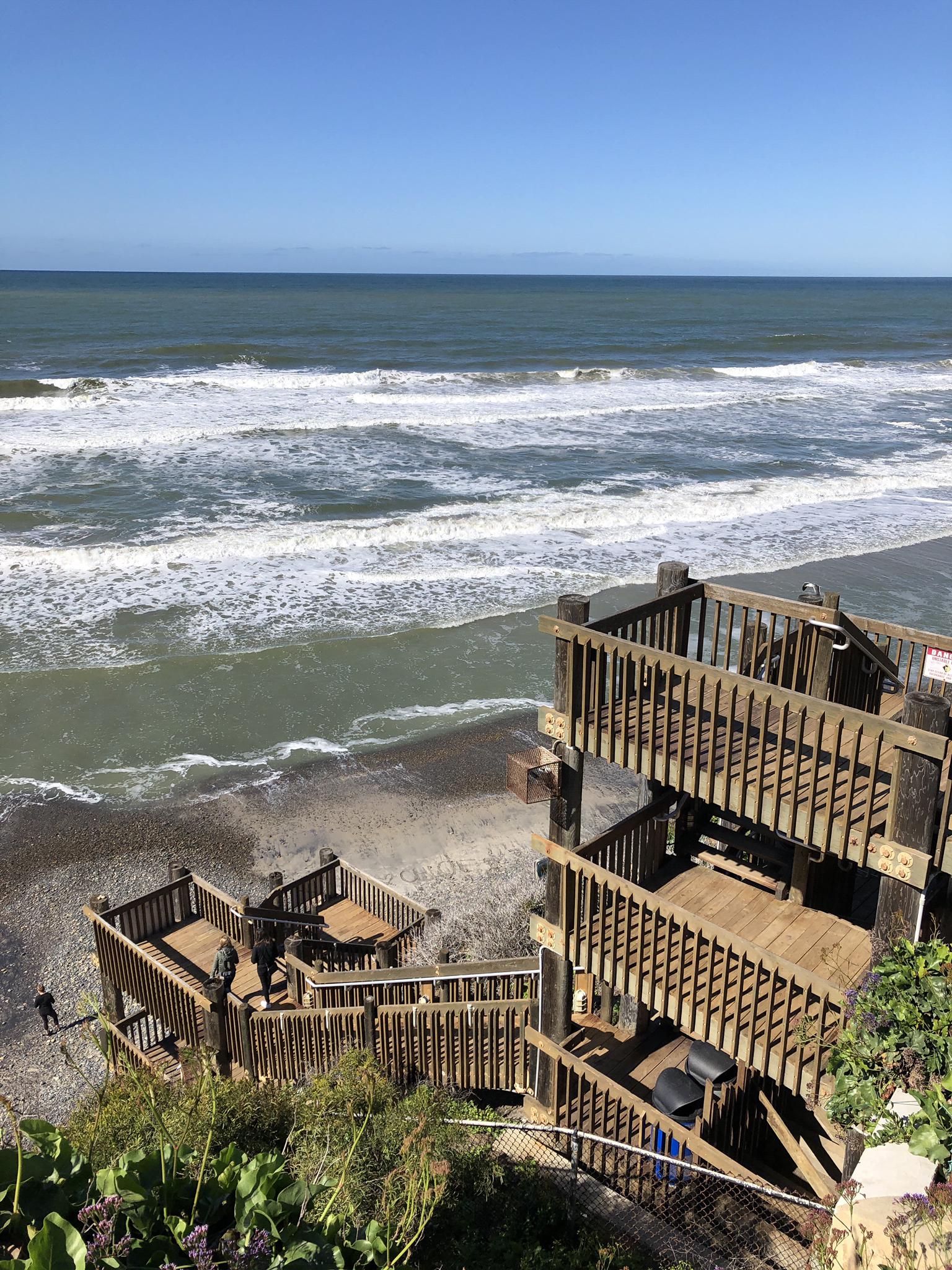 Wooden stairs descend a cliff to a sunny beach with crashing ocean waves.
