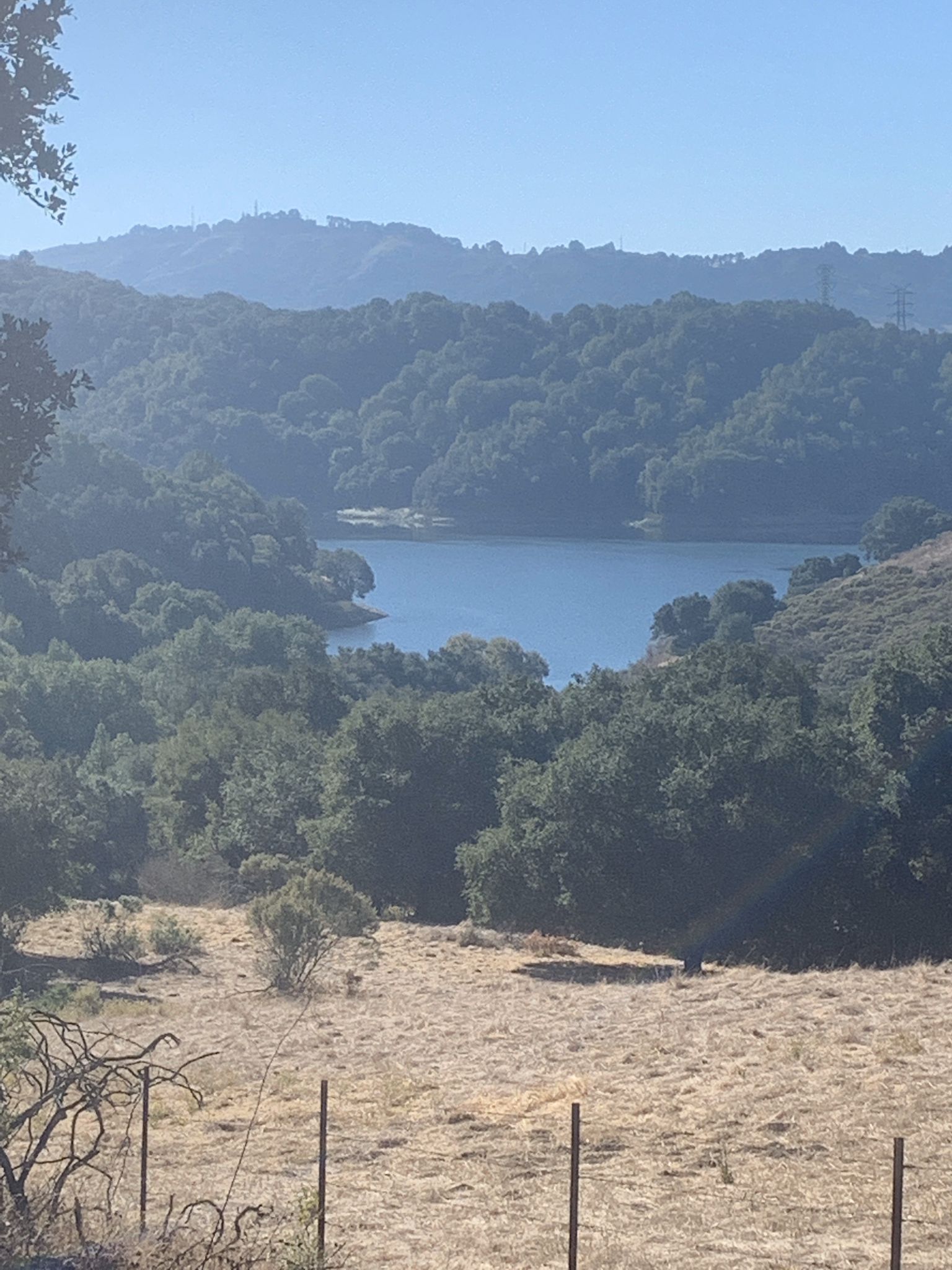 A serene lake nestled among green hills under a bright, hazy sky. - Three Bears Loop