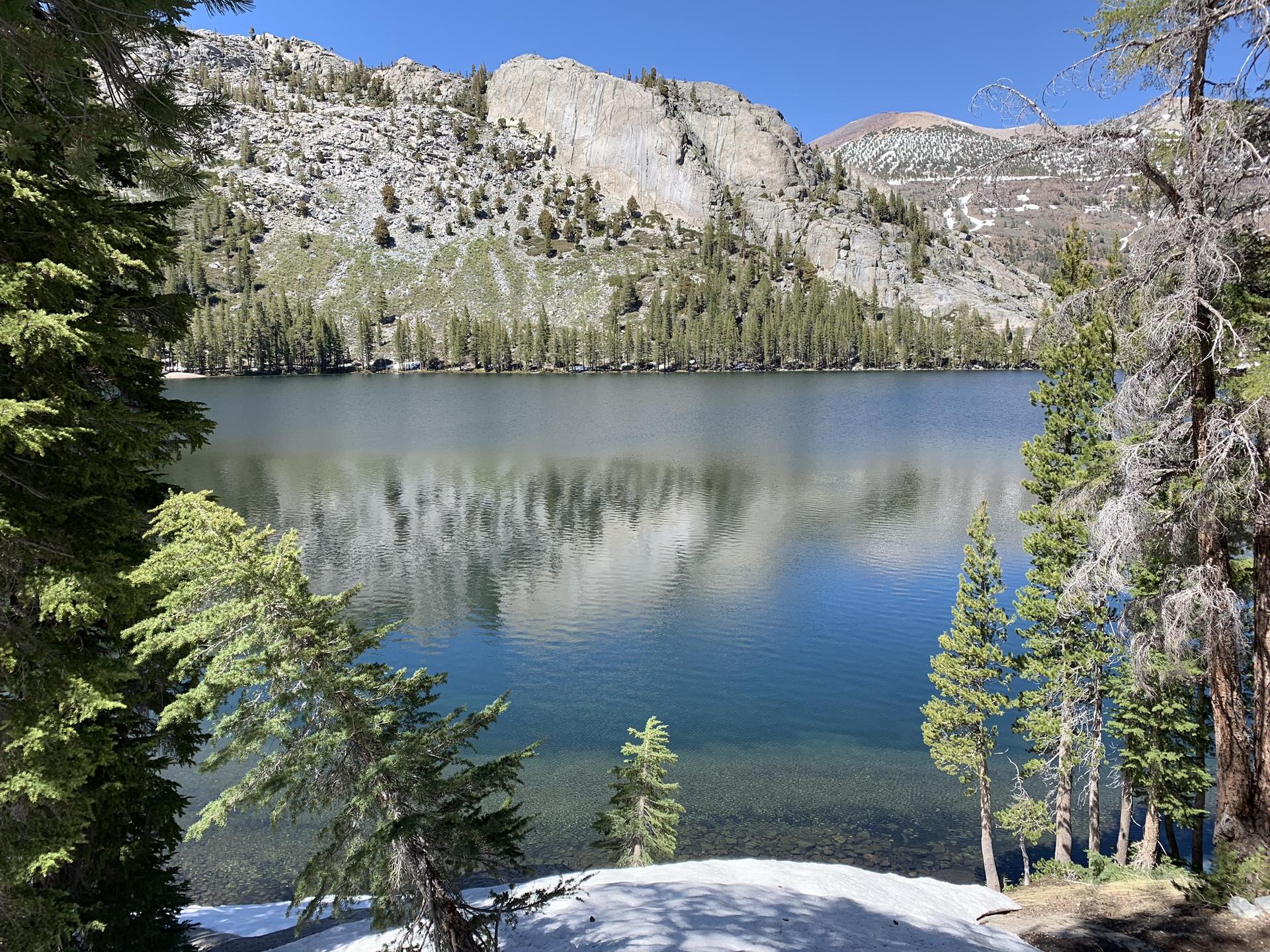 Clear mountain lake framed by pine trees, with rocky, snow-dusted peaks.