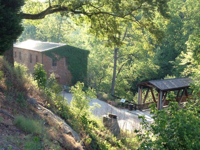 A brick building and covered bridge nestled in a sunny, green, wooded hillside.