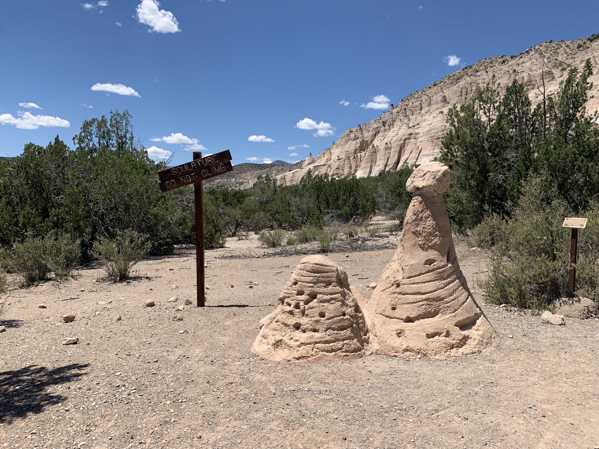 Two small hoodoos and a wooden sign stand on a dirt path under a blue sky.