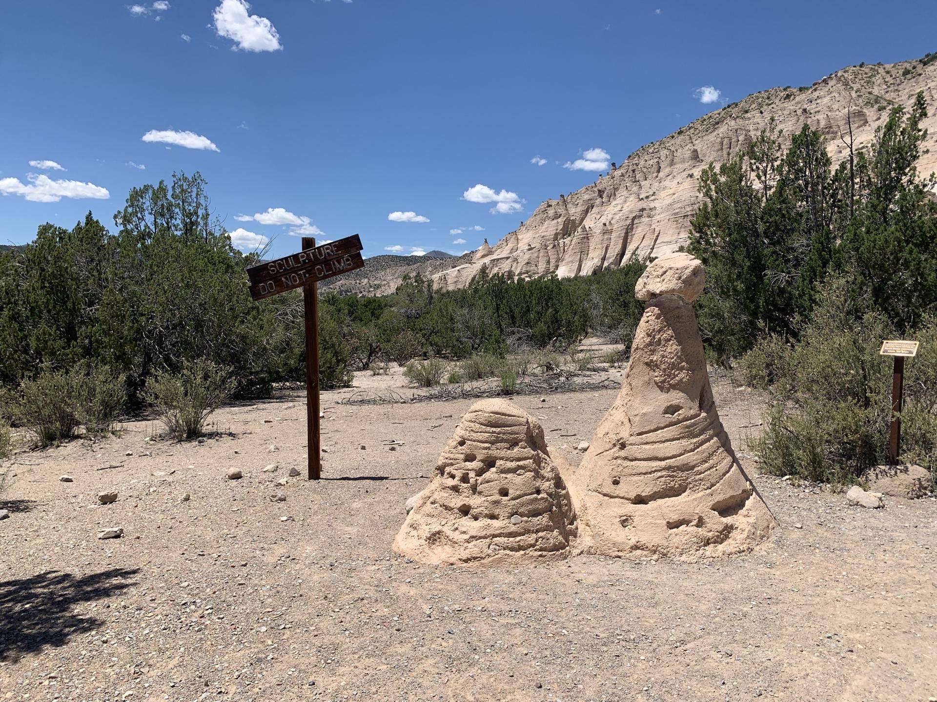 Two small hoodoos and a wooden sign stand on a dirt path under a blue sky.