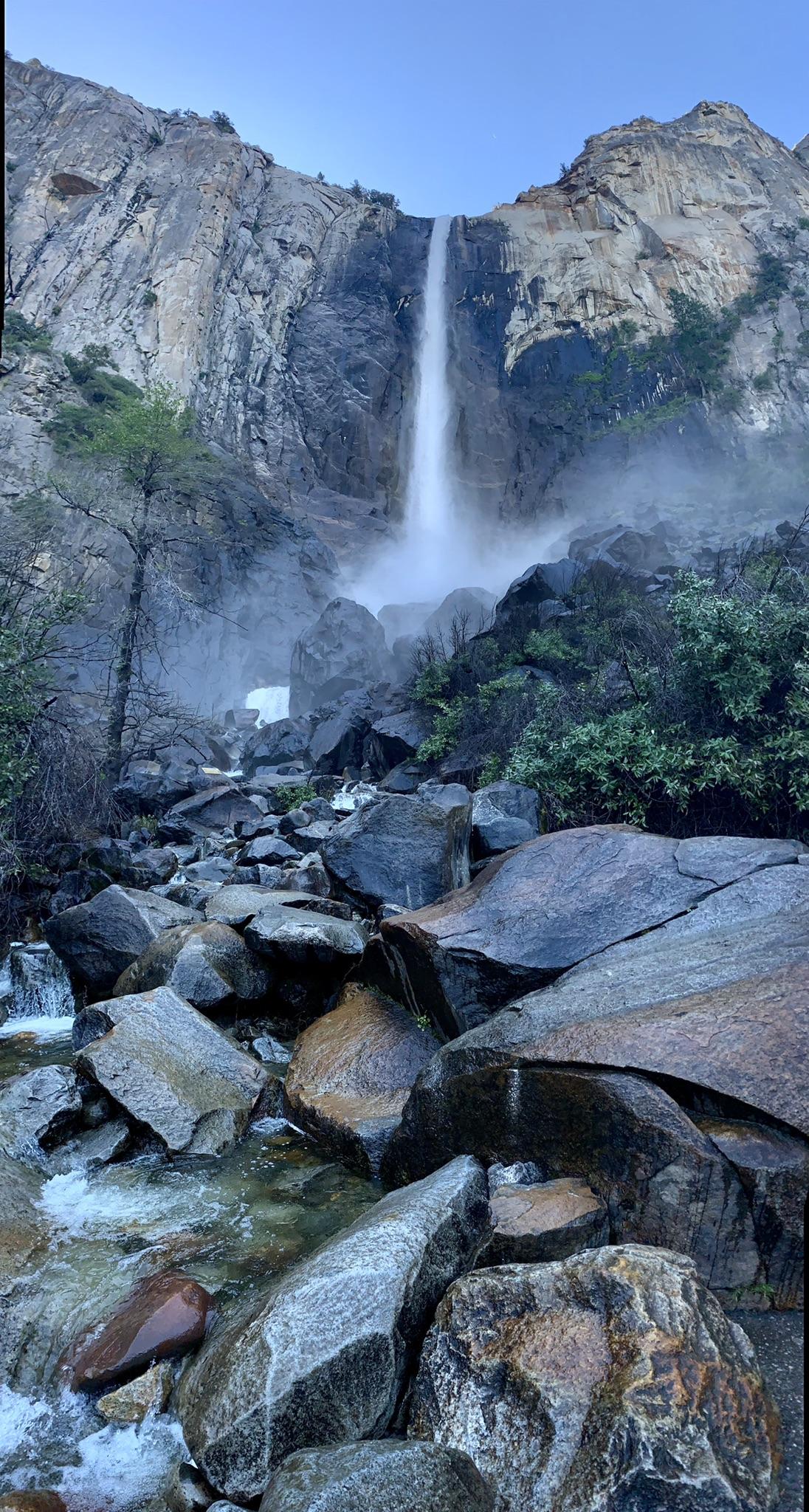 Tall waterfall cascades down a rocky cliff into a misty, boulder-filled stream.