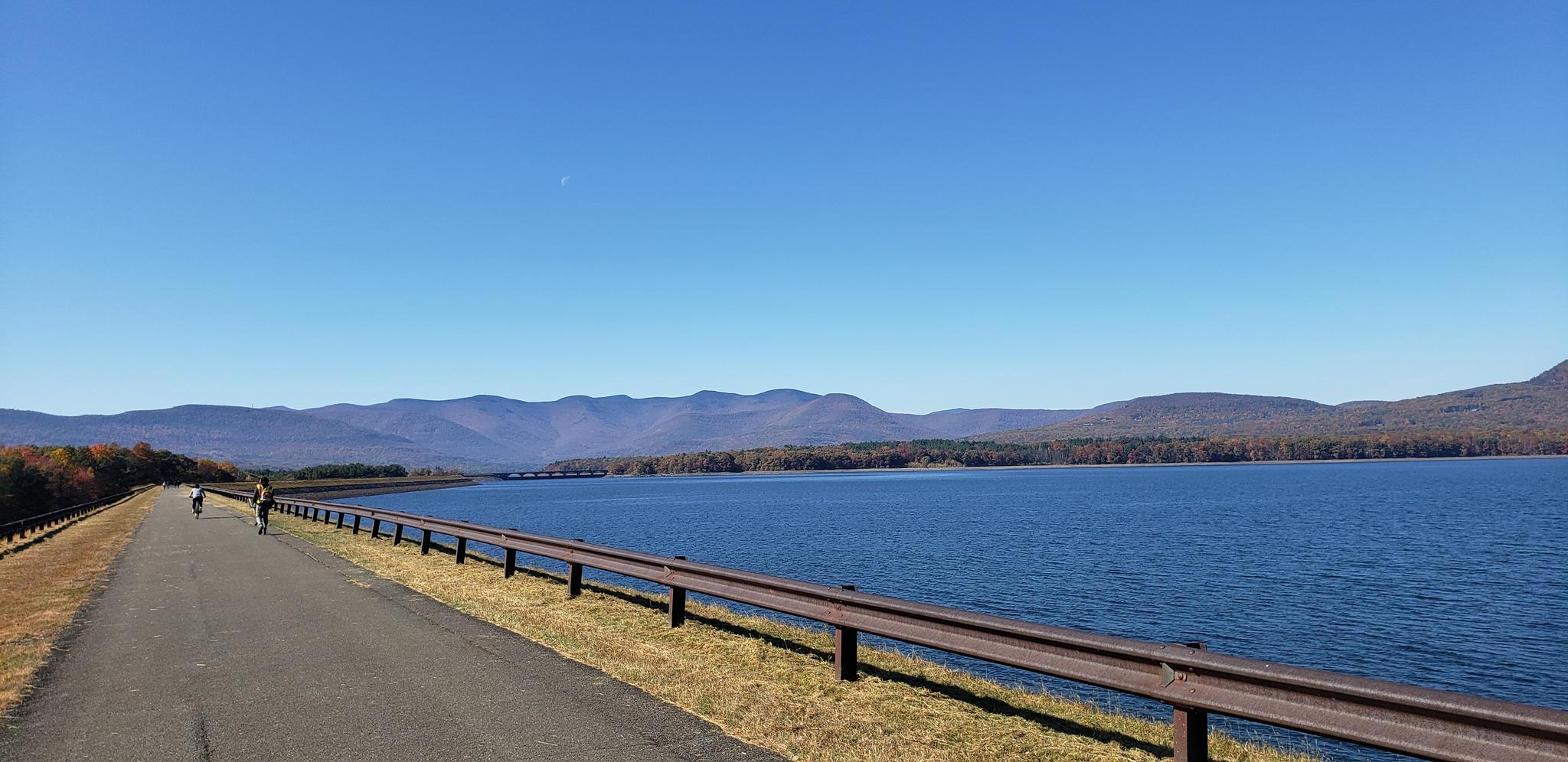 Paved road beside a vast lake with distant mountains, two cyclists, sunny day.