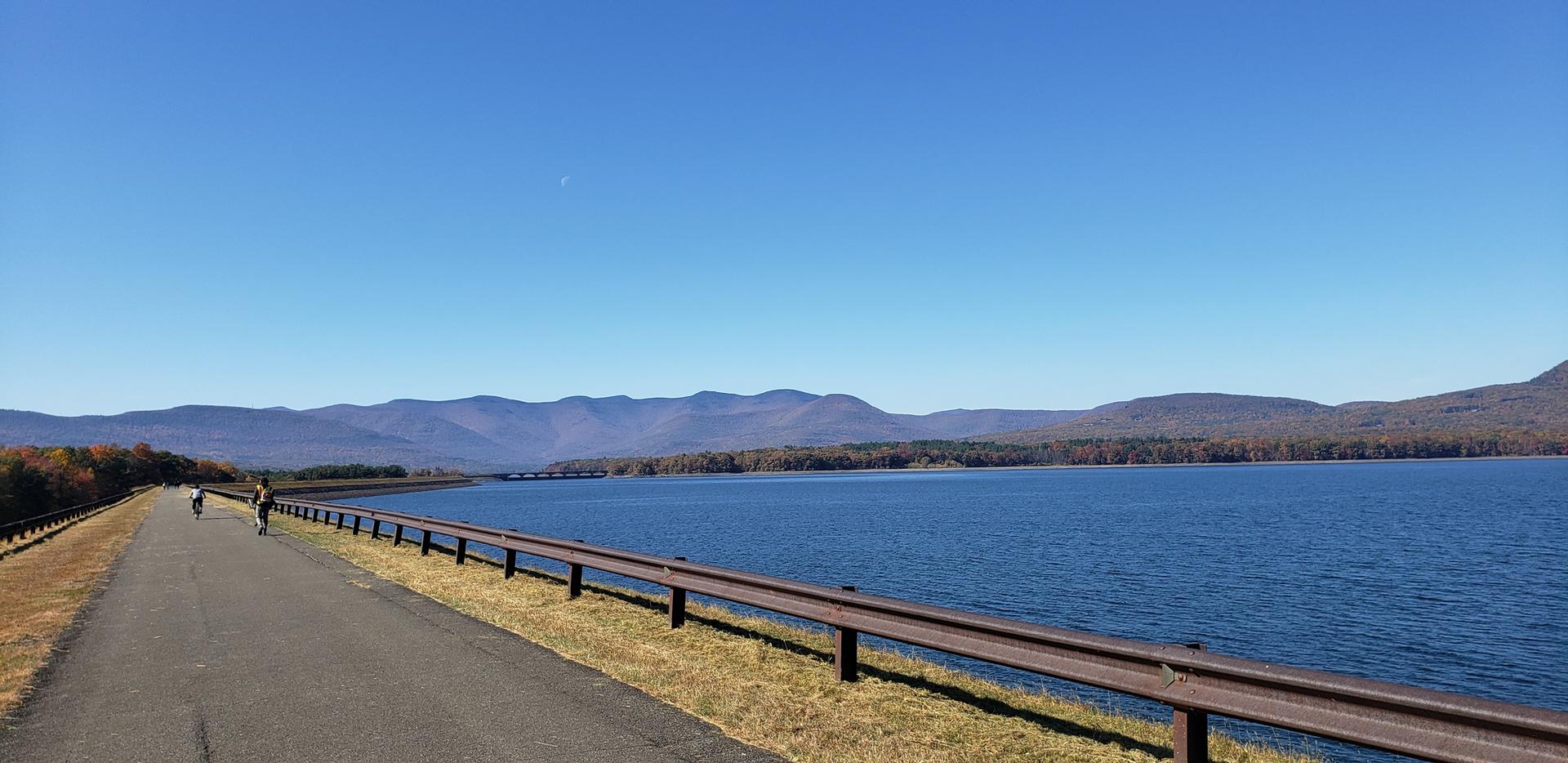 Paved road beside a vast lake with distant mountains, two cyclists, sunny day.