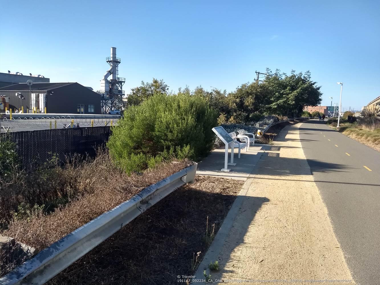 Paved path with benches next to industrial structures and bushes on a sunny day.