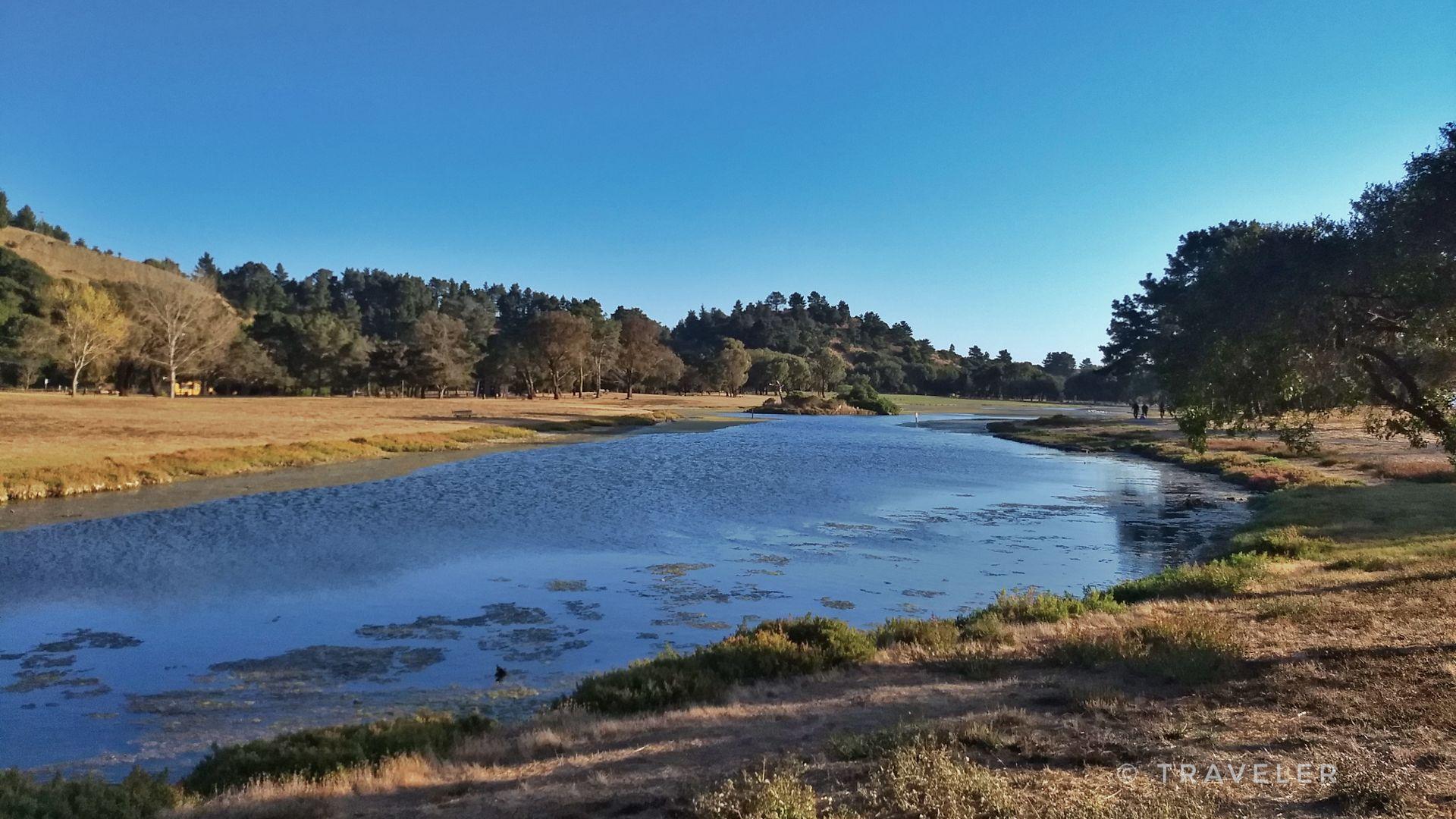 Serene lake surrounded by grassy hills and trees under a clear blue sky.