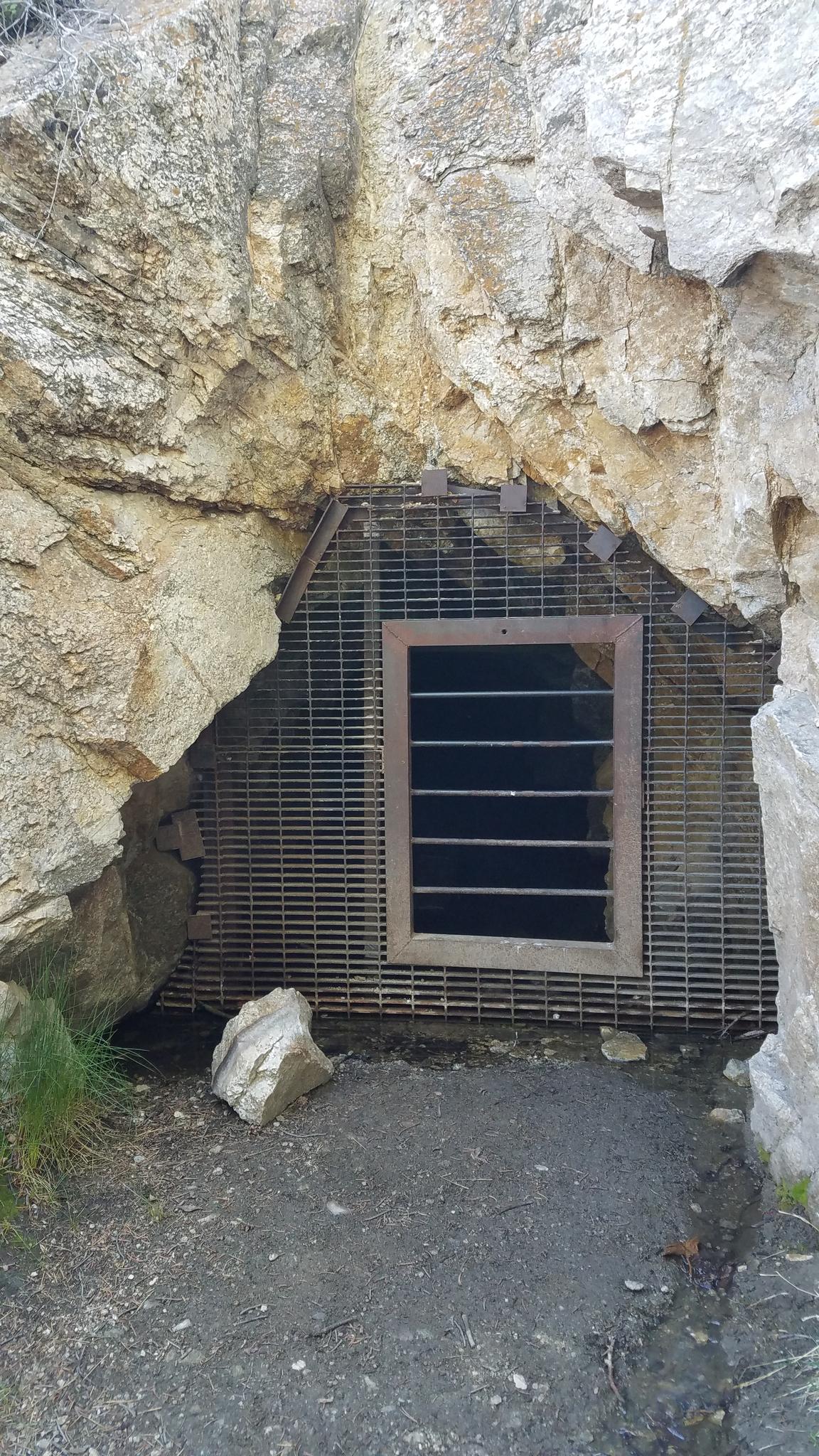 A dark cave entrance covered by a metal grate in a rocky cliff.