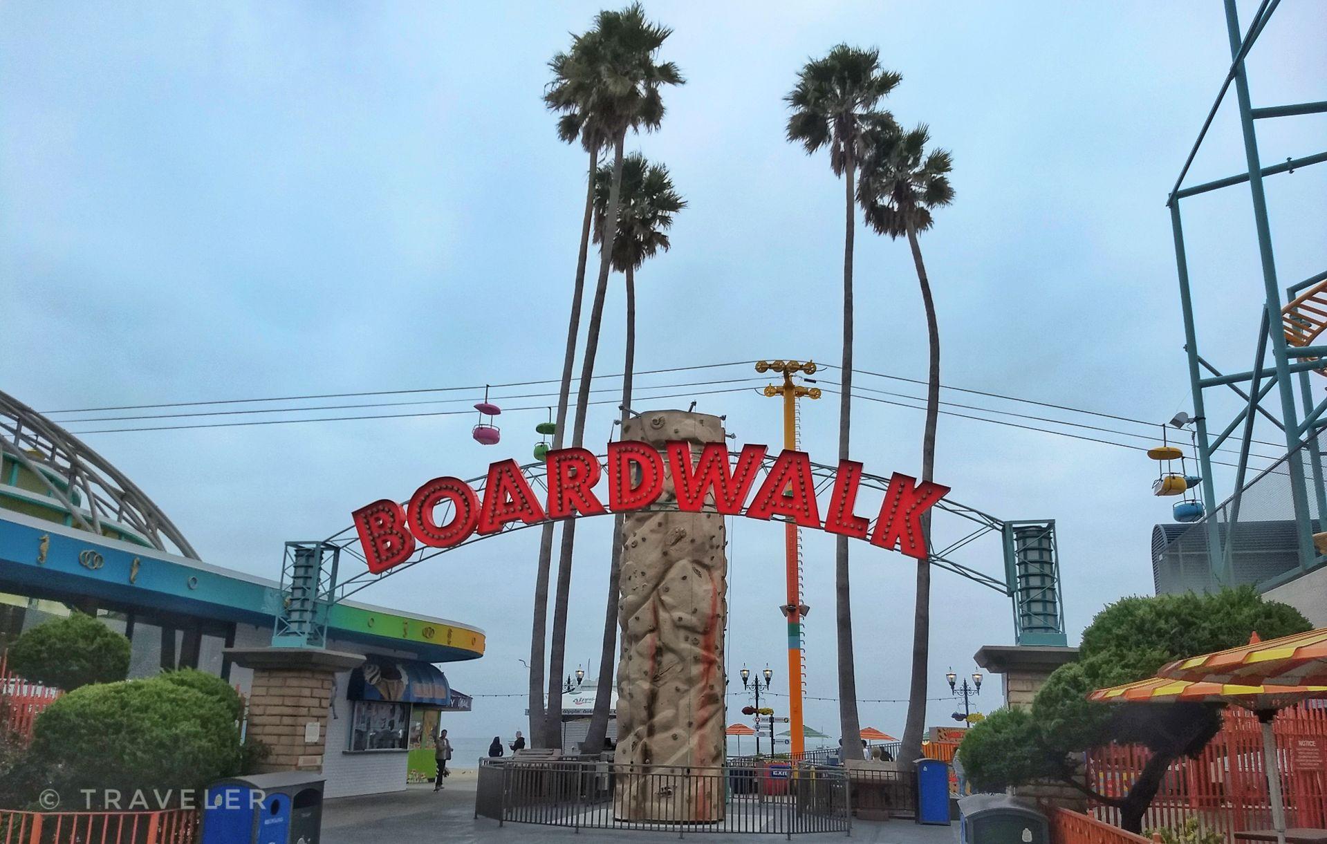Entrance to a boardwalk with a large sign, palm trees, and amusement rides.