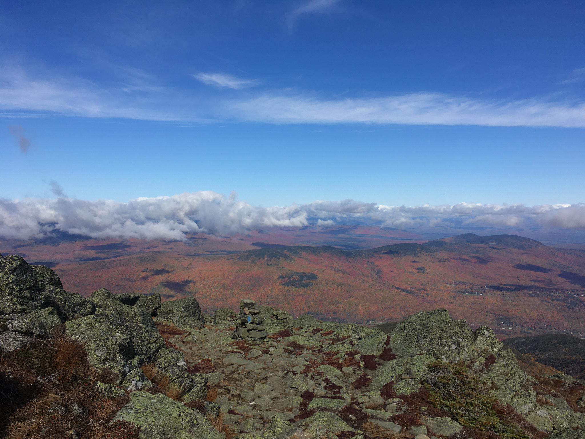 Rocky mountain summit overlooking a vast autumn landscape with clouds below.