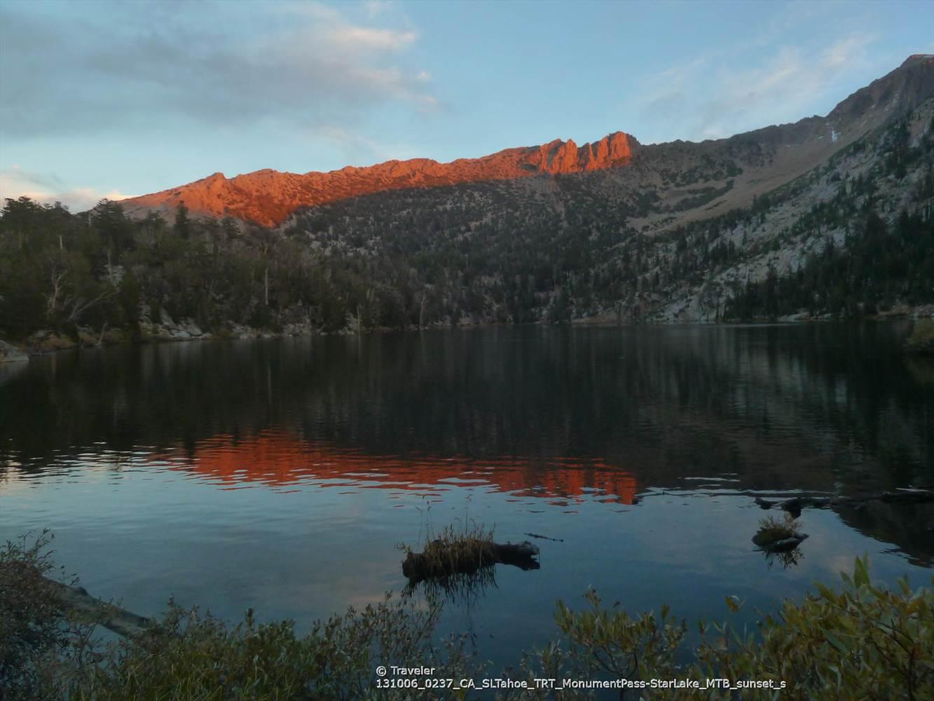 A serene mountain lake reflects orange sunset light on distant peaks.