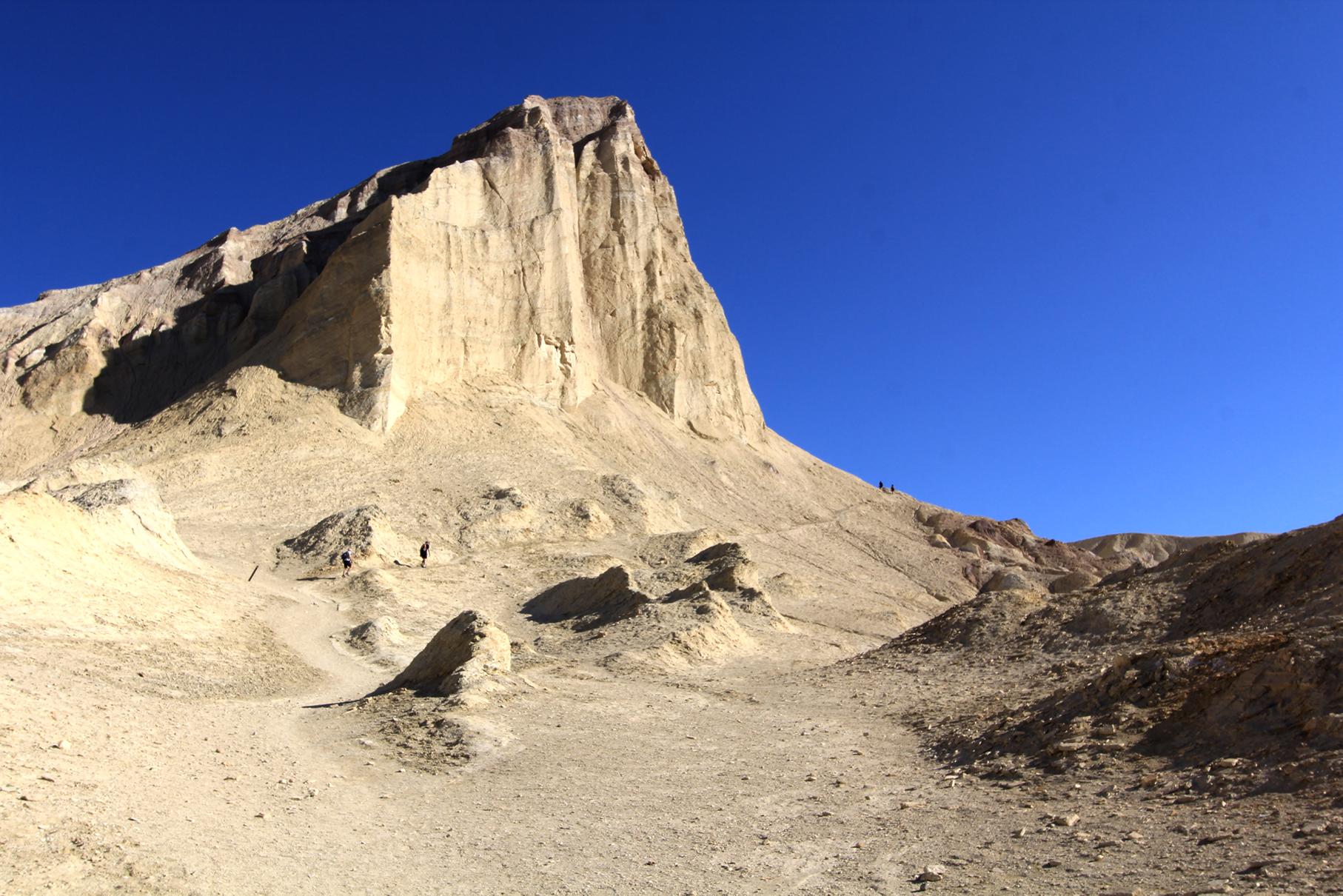 Two small figures walk near a tall, light-colored rock formation under a bright blue sky.